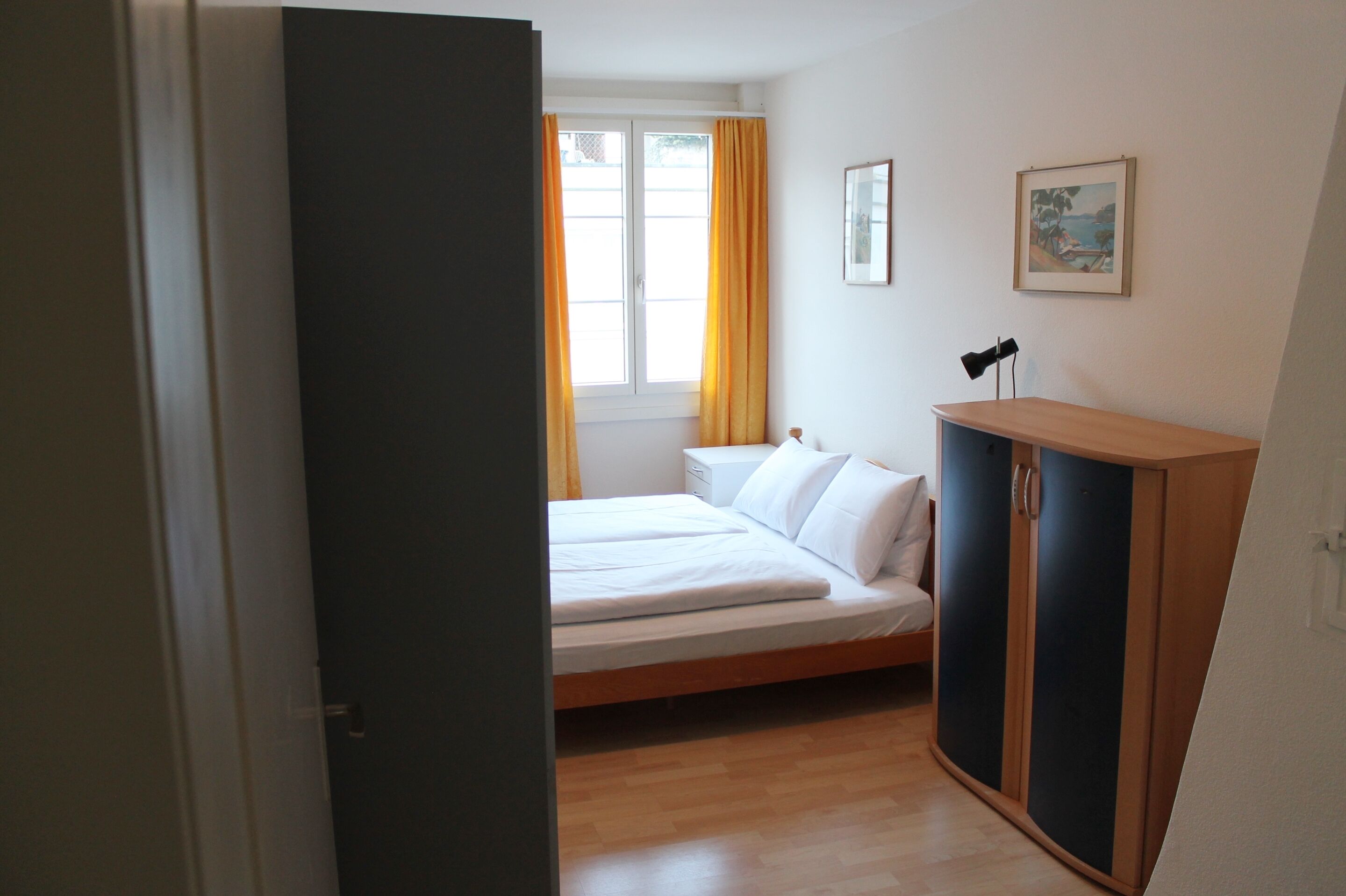 A bedroom with a bed, a wardrobe, a bedside table, a window with curtains, and a wall-mounted picture.