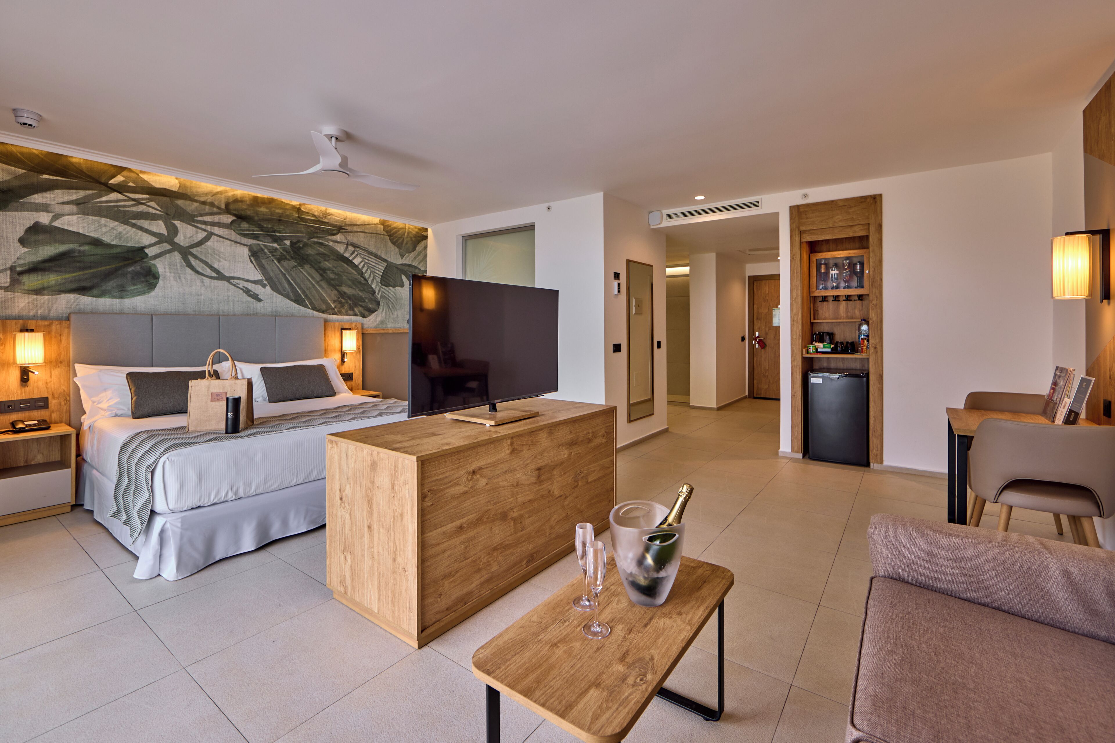 A modern hotel room with a bed, a wooden desk, a television, and a minibar.