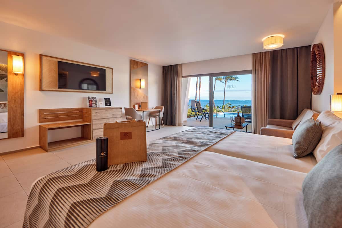 A modern hotel room with a large bed, a desk, a chair, a sofa, and a balcony with a view of the sea.
