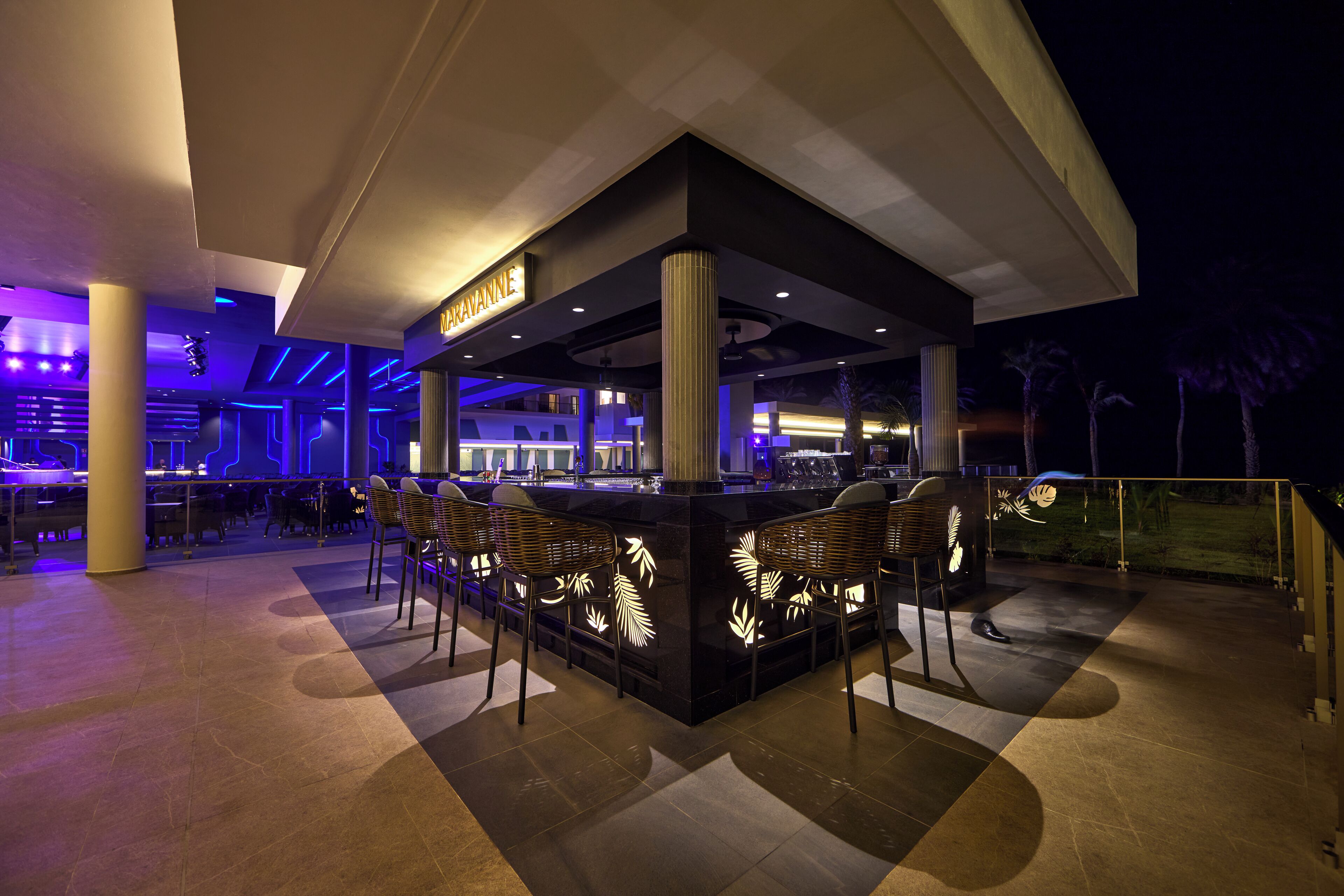 Zona de bar al aire libre con diseño moderno, que cuenta con una barra central con asientos y patrones de hojas iluminadas.