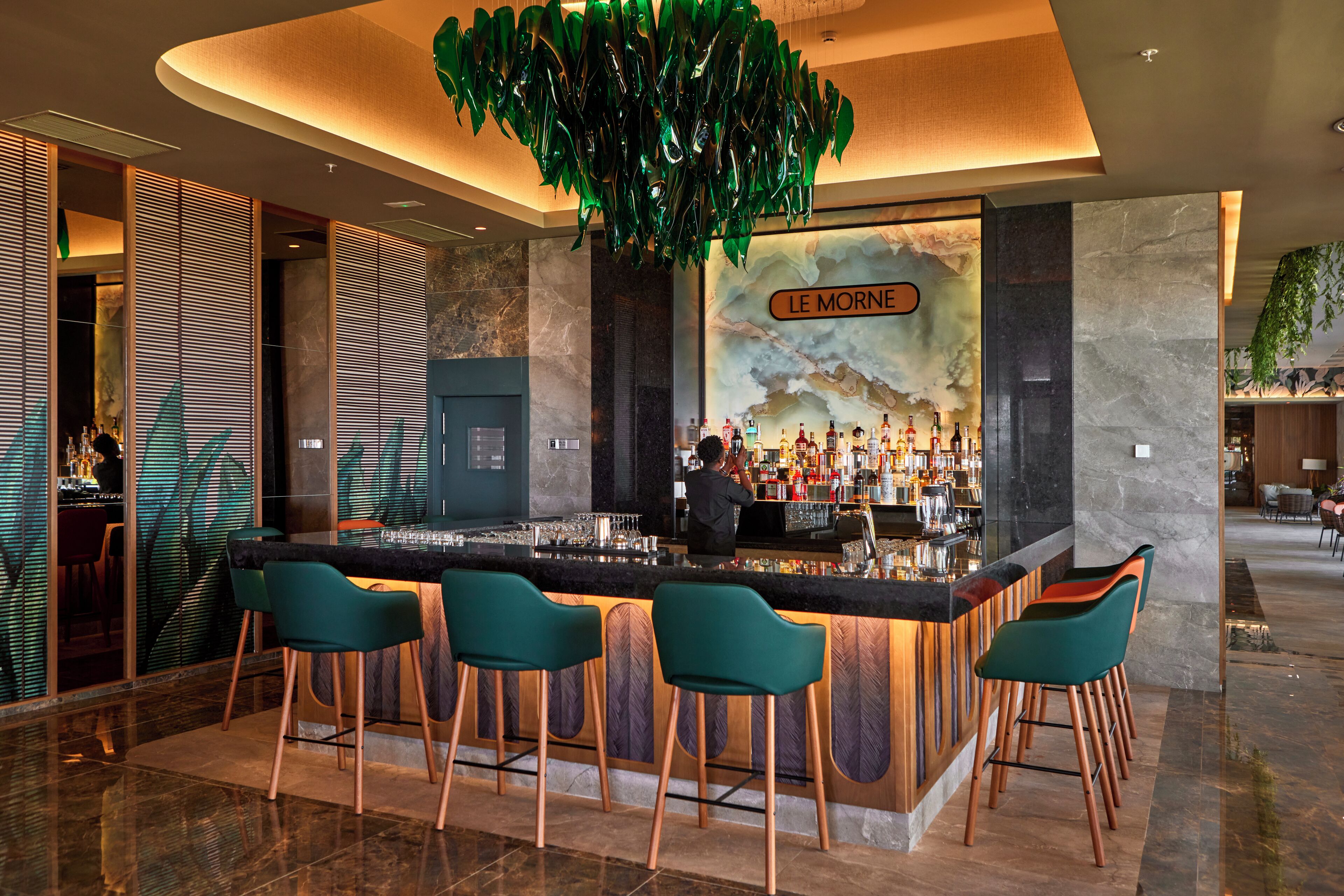 A bar area with a modern design, featuring a bar counter, green bar stools, and a bartender.