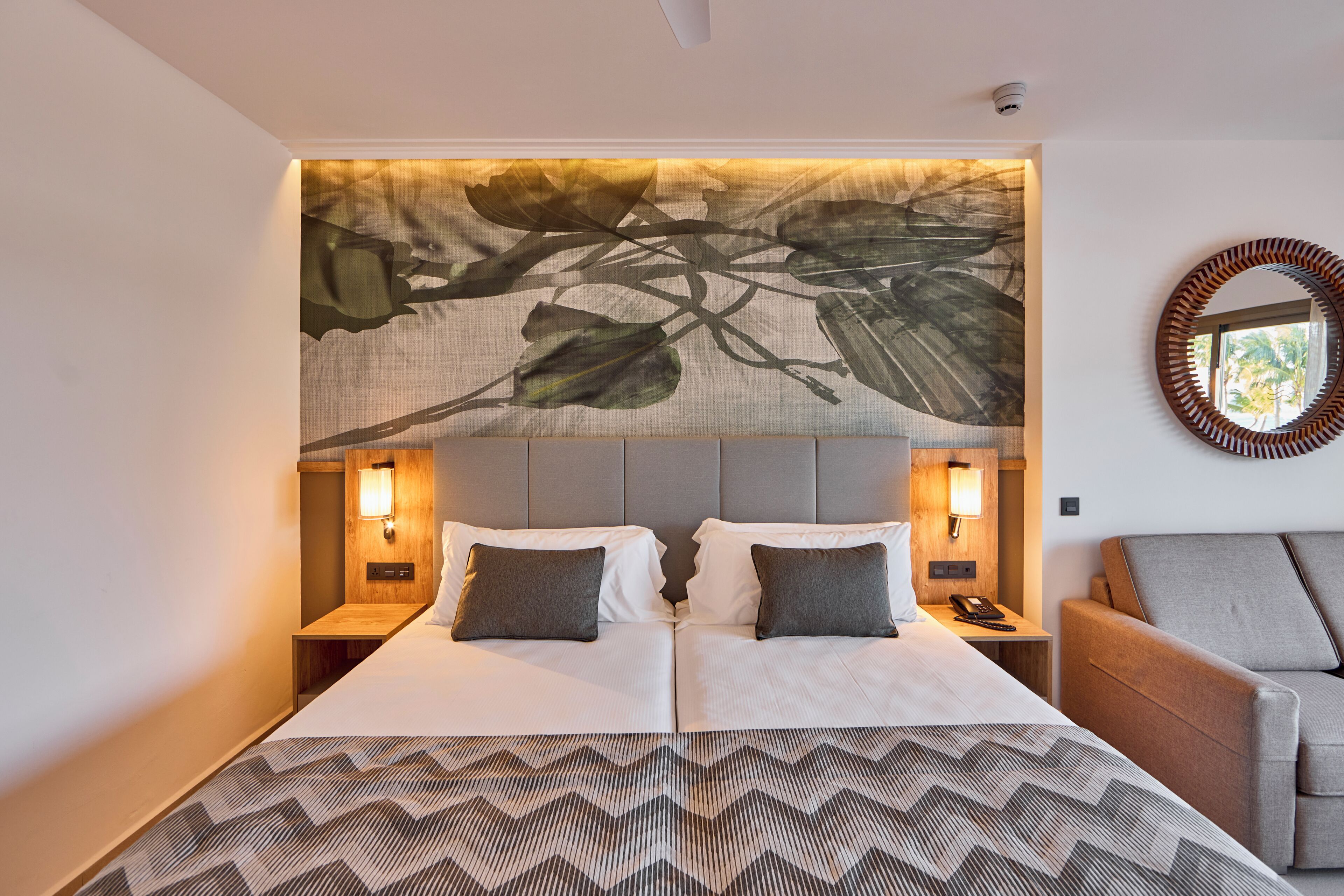 A double bed with a patterned bedspread, two bedside tables with lamps, a wall-mounted mirror, and a view of the outdoors.