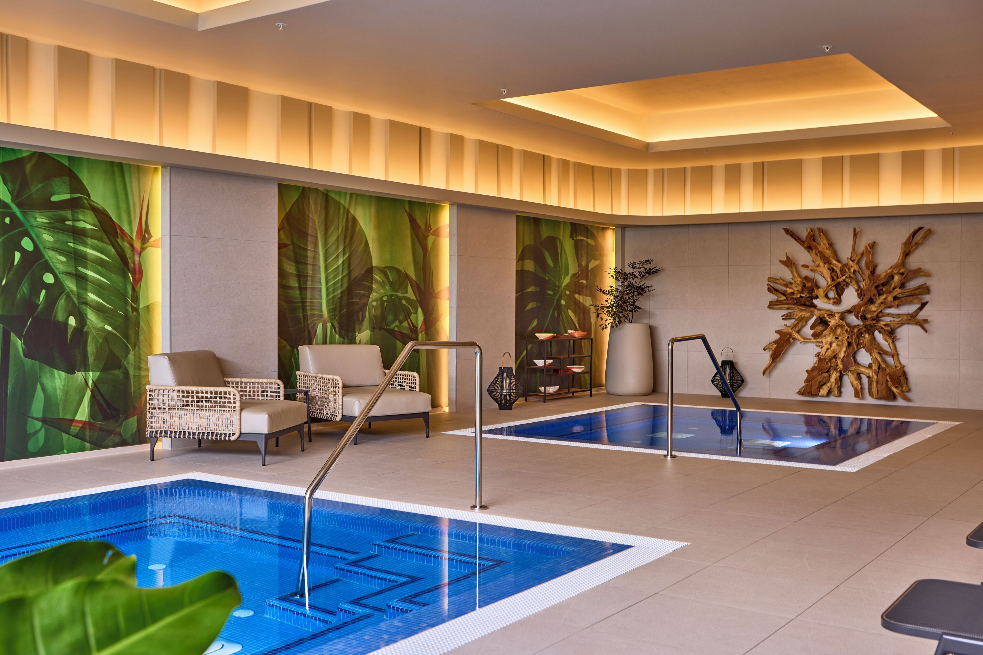 A modern indoor swimming pool with two separate sections, surrounded by lounge chairs and a decorative wall with a tropical mural.