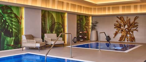 A modern indoor swimming pool with two separate sections, surrounded by lounge chairs and a decorative wall with a tropical mural.