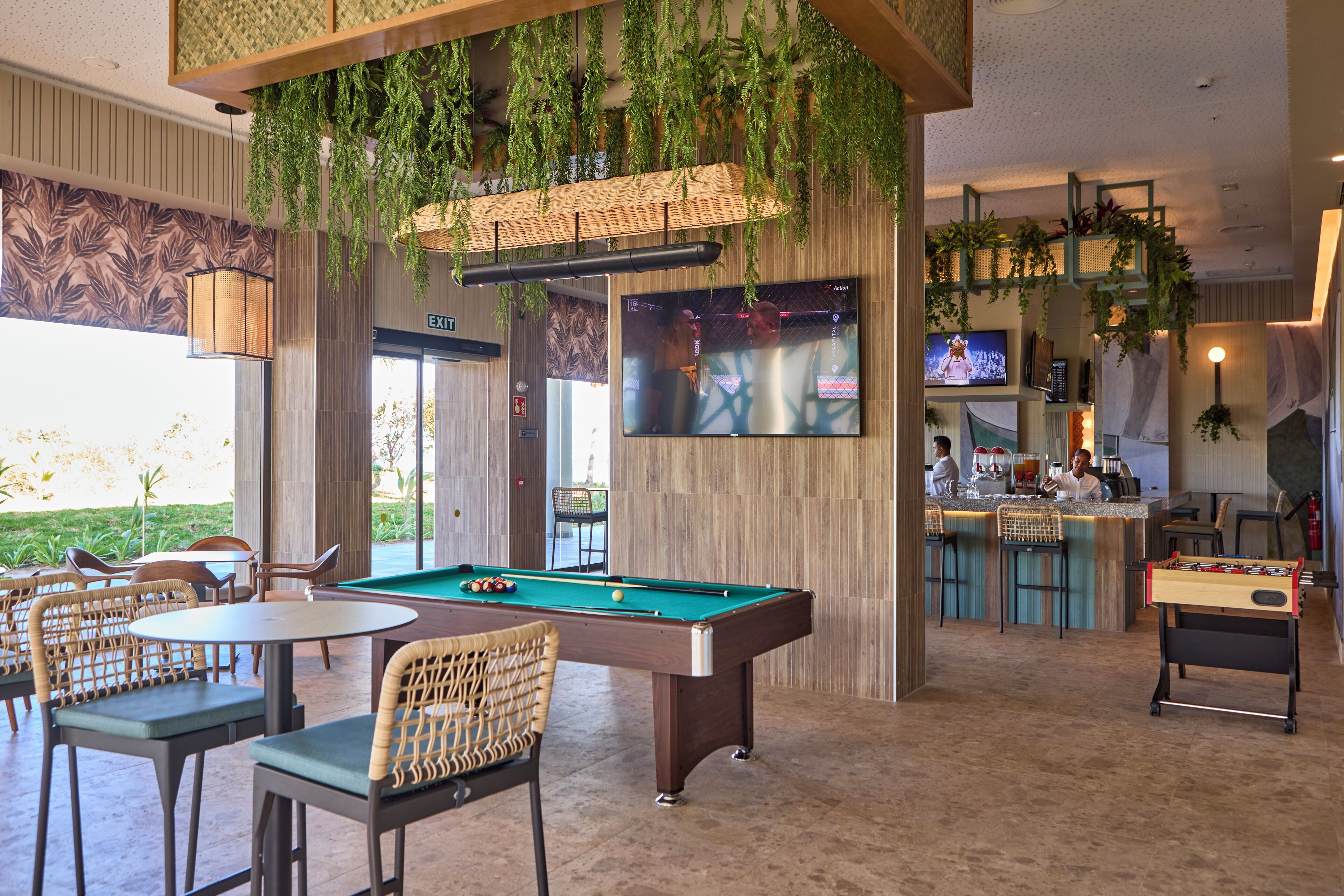 A pool table, bar area, seating, and hanging plants in a room with a wooden wall and a TV screen.