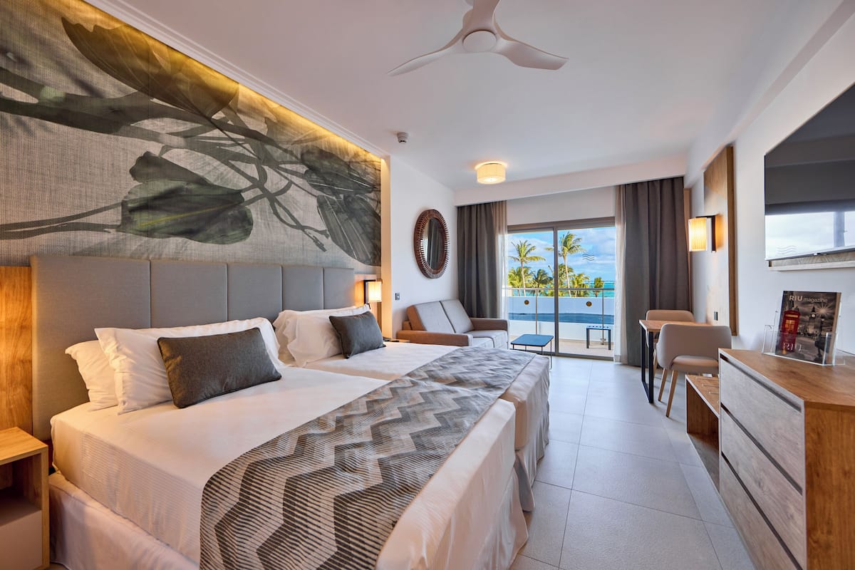 A hotel room with two beds, a desk, and a view of palm trees.