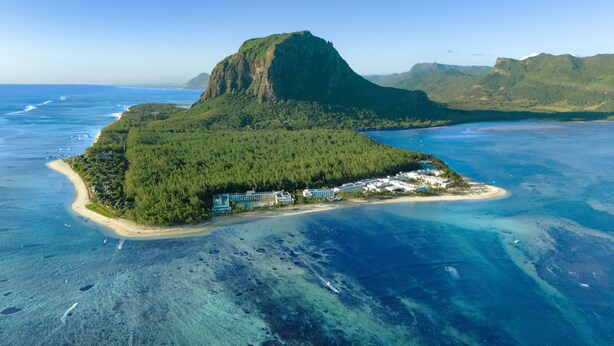 An aerial view of a coastal resort with a prominent mountain, clear blue waters, and a small peninsula with buildings.