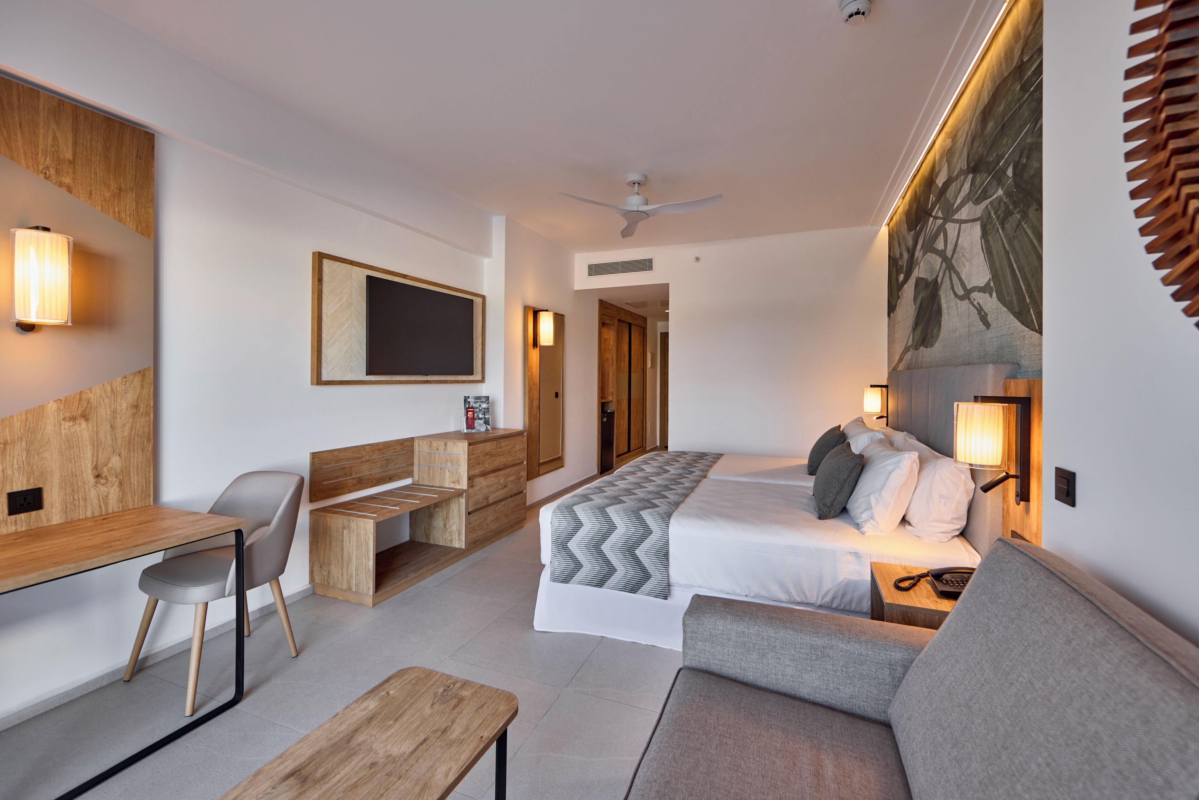 A modern hotel room with a bed, a desk, a chair, a sofa, and a wooden headboard.