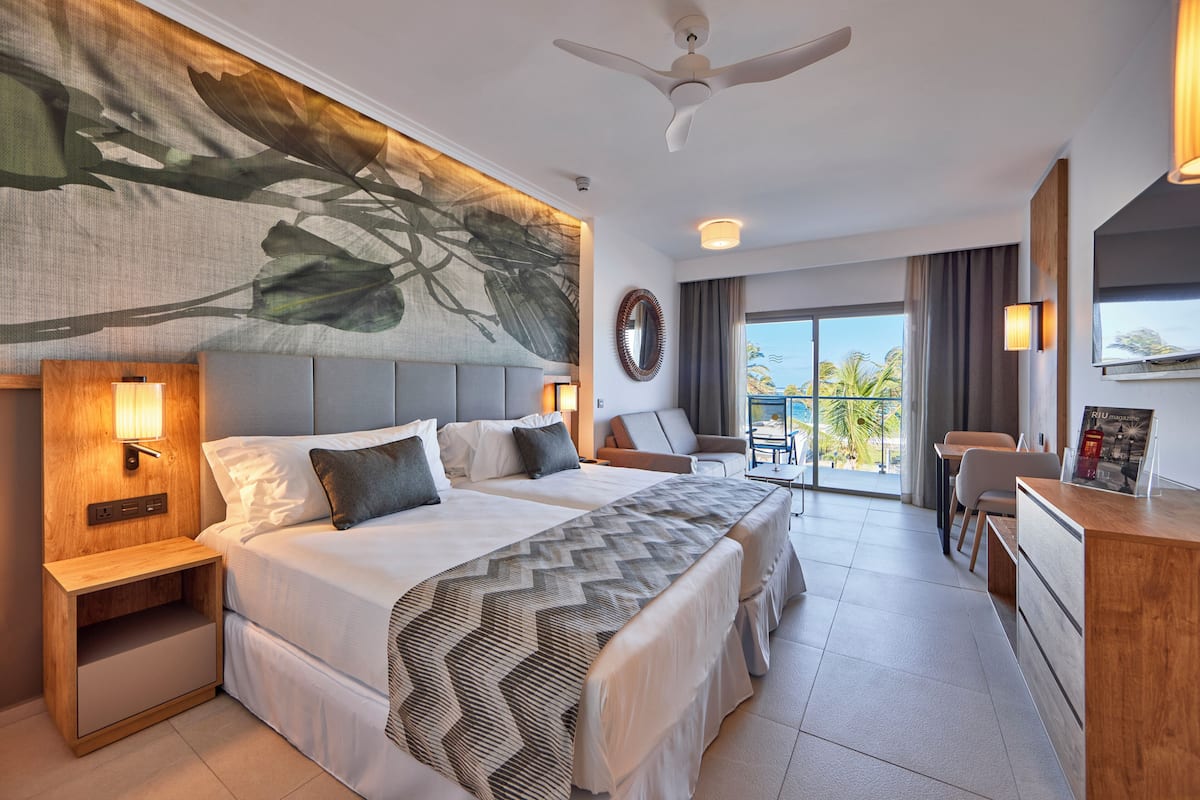 A hotel room with a large bed, two bedside tables with lamps, a ceiling fan, a view of the outdoors, and a desk with a chair.