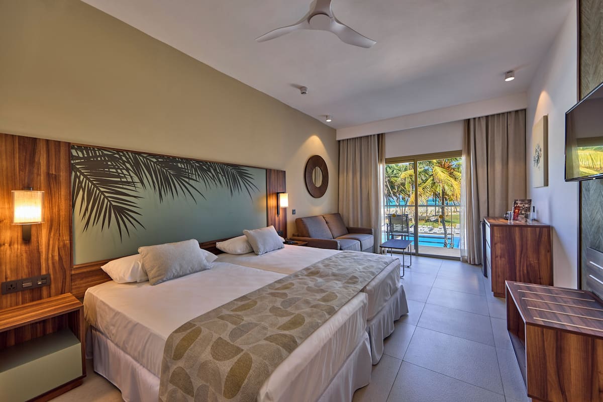 A hotel room with a large bed, a desk, a chair, a TV, and a view of the pool.