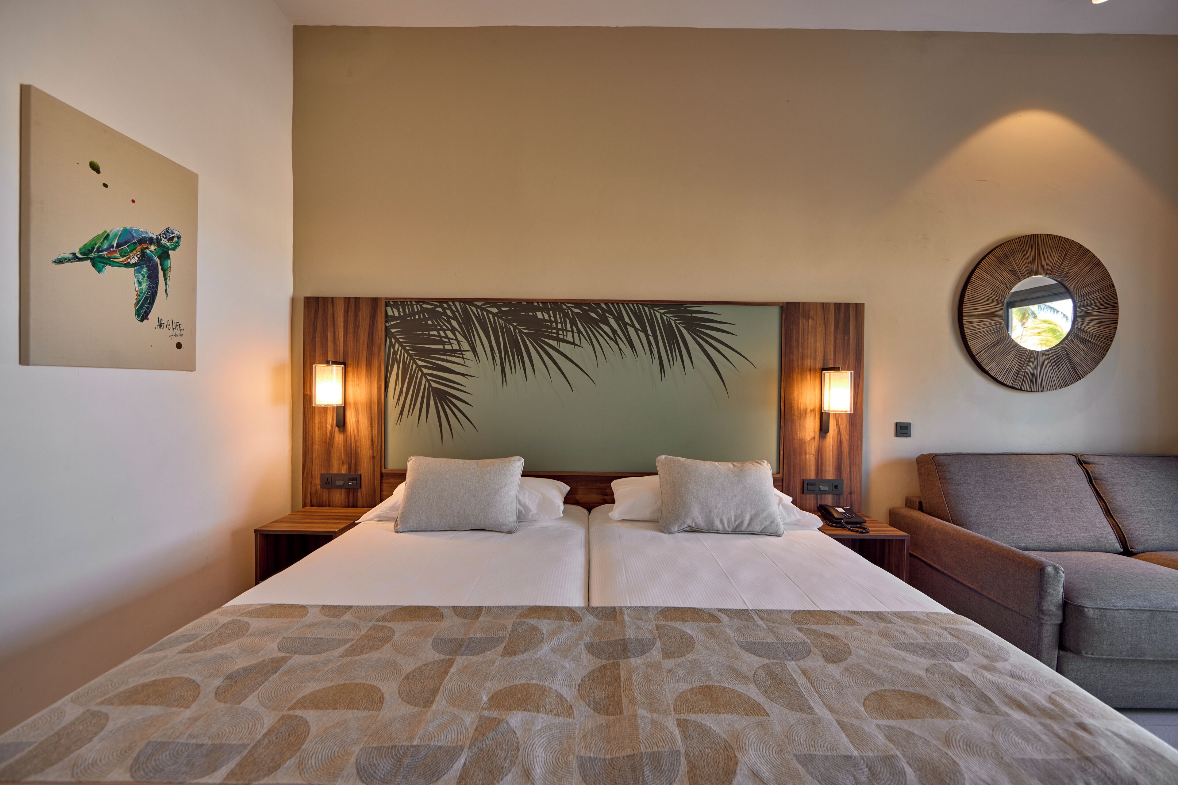 Une chambre d’hôtel avec un grand lit, deux tables de chevet, un canapé, un miroir fixé au mur et une œuvre d’art sur le thème des tropiques.