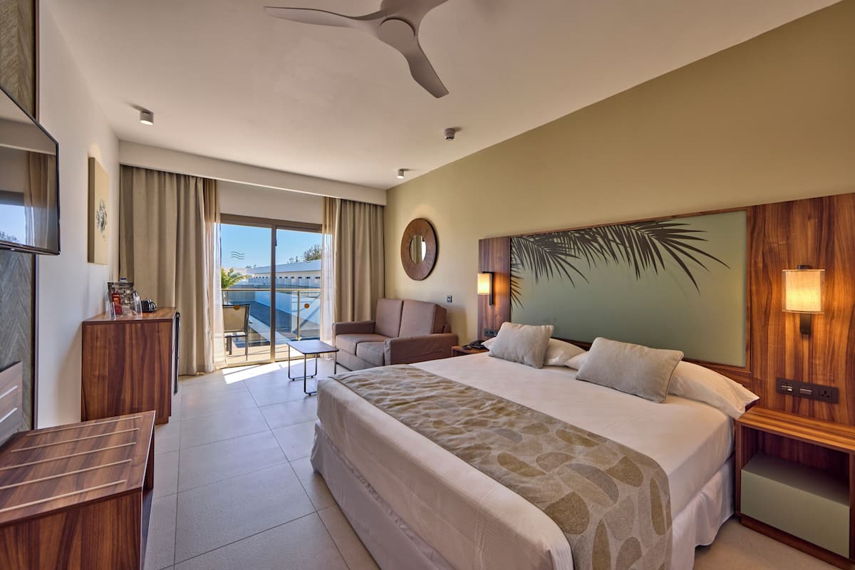 A hotel room with a large bed, a desk, a chair, a small table, a ceiling fan, and a view of the outdoors.