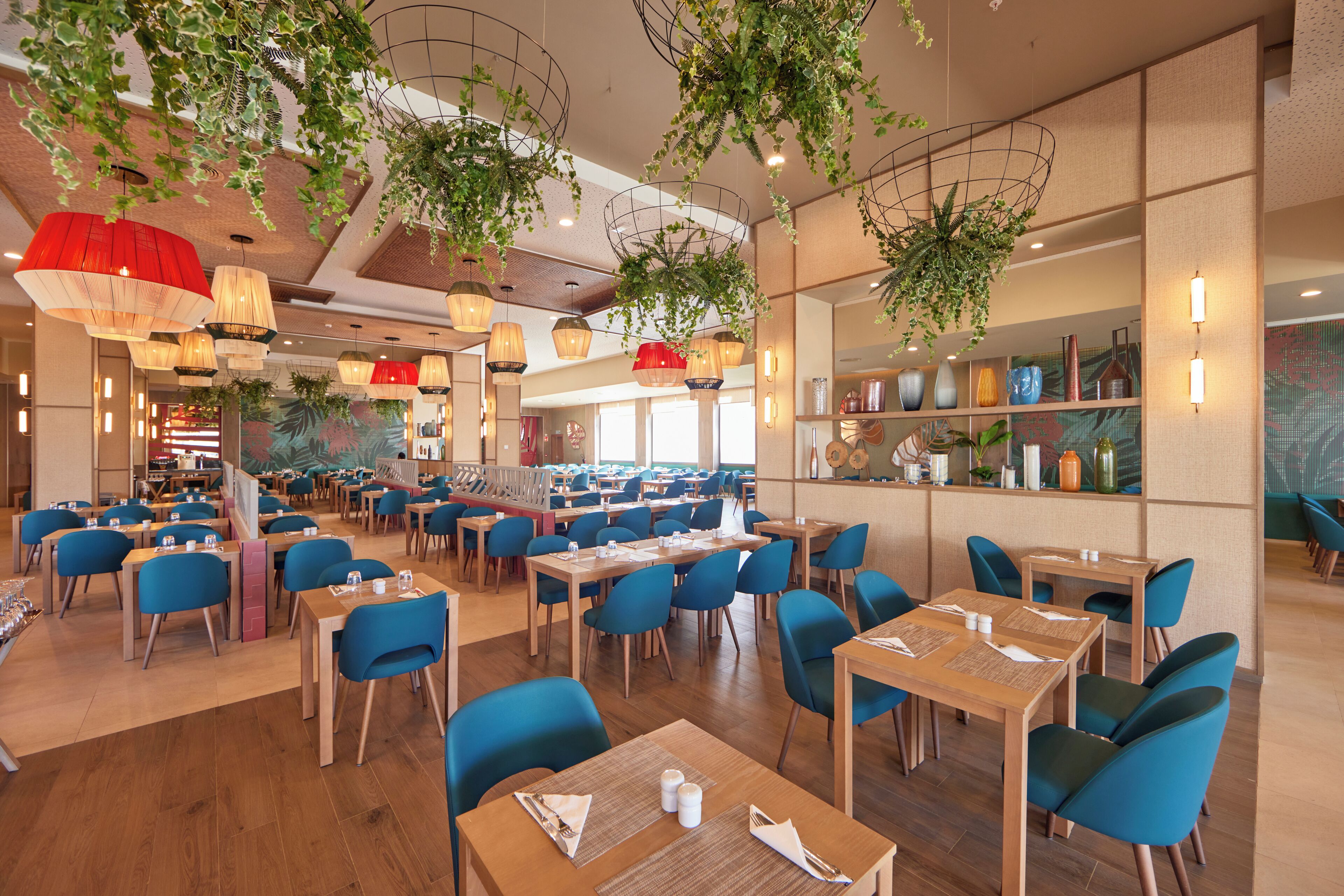 A spacious restaurant with multiple tables and blue chairs, featuring hanging plants and pendant lights.