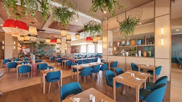 A spacious restaurant with multiple tables and blue chairs, featuring hanging plants and pendant lights.