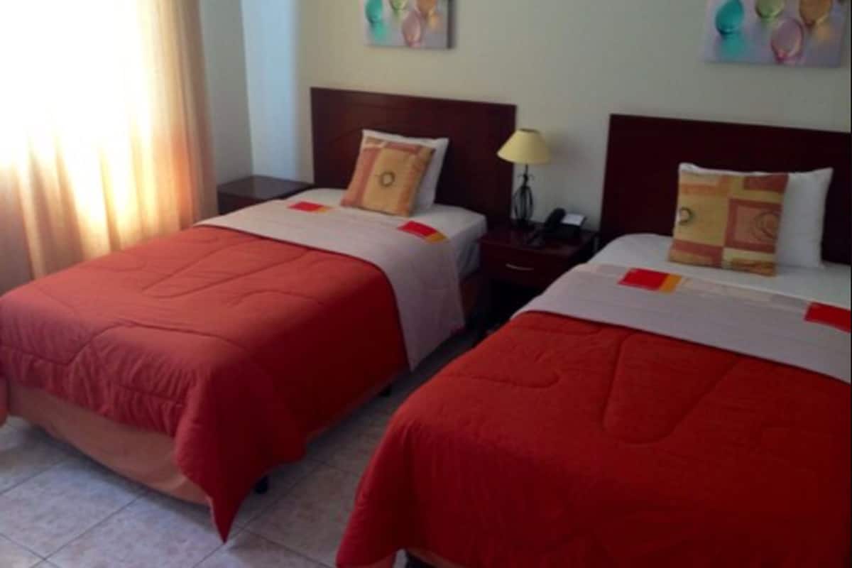 Une chambre d’hôtel avec deux lits, chacun recouvert d’une courtepointe rouge et agrémenté d’oreillers assortis. Une table de chevet avec une lampe et un téléphone se trouve entre les deux lits. La chambre est dotée d’une fenêtre avec des rideaux, et on peut apercevoir des œuvres d’art sur le mur.