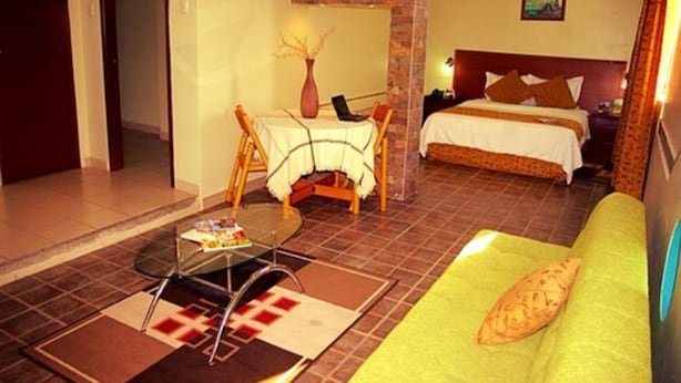 Habitación de hotel con una cama, un sofá verde, una mesa de comedor con sillas y una mesita con una computadora portátil y un florero.