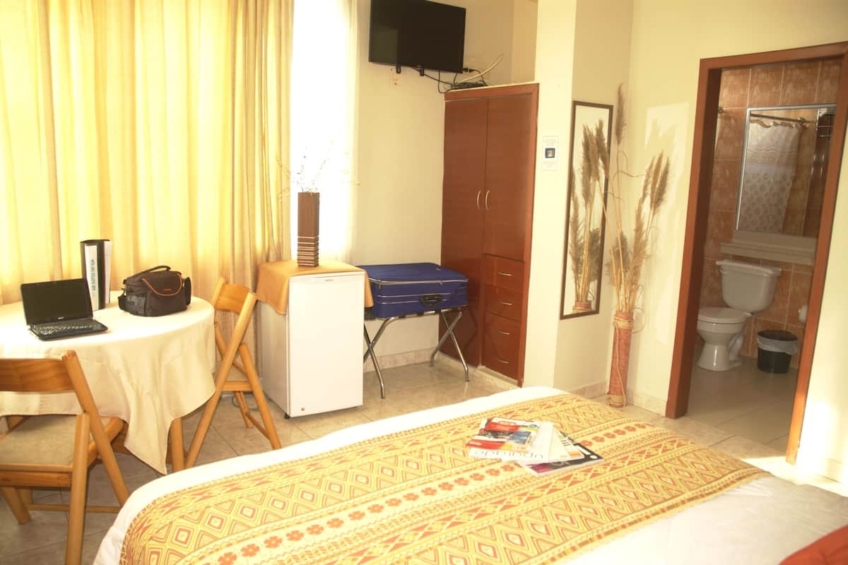 Une chambre d’hôtel comprenant un lit, un bureau avec un ordinateur portable, une valise, une armoire et une salle de bain avec des toilettes et une douche.