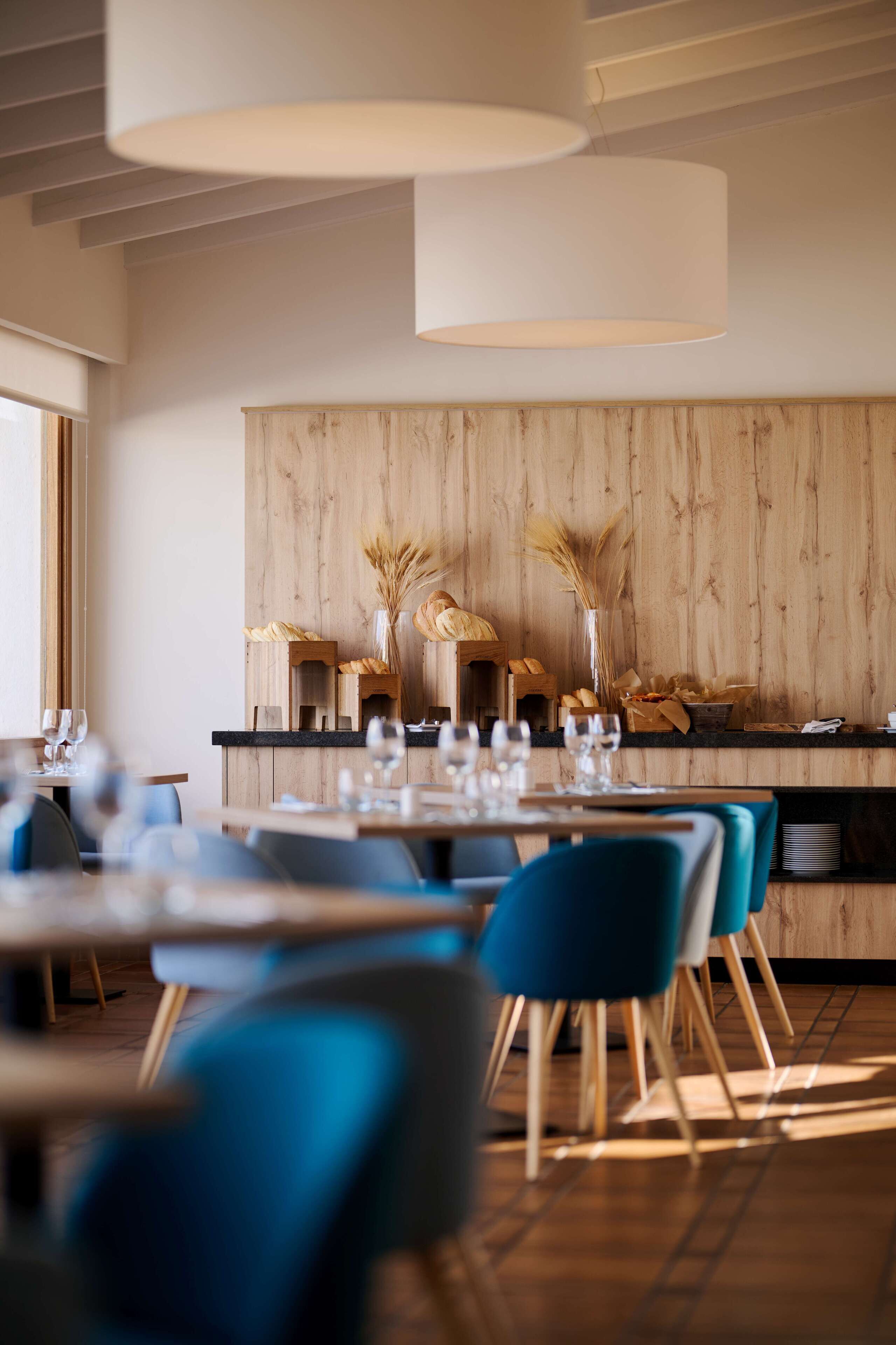 Interior de un restaurante moderno con paneles de madera, sillas azules y una barra central con pan y flores.