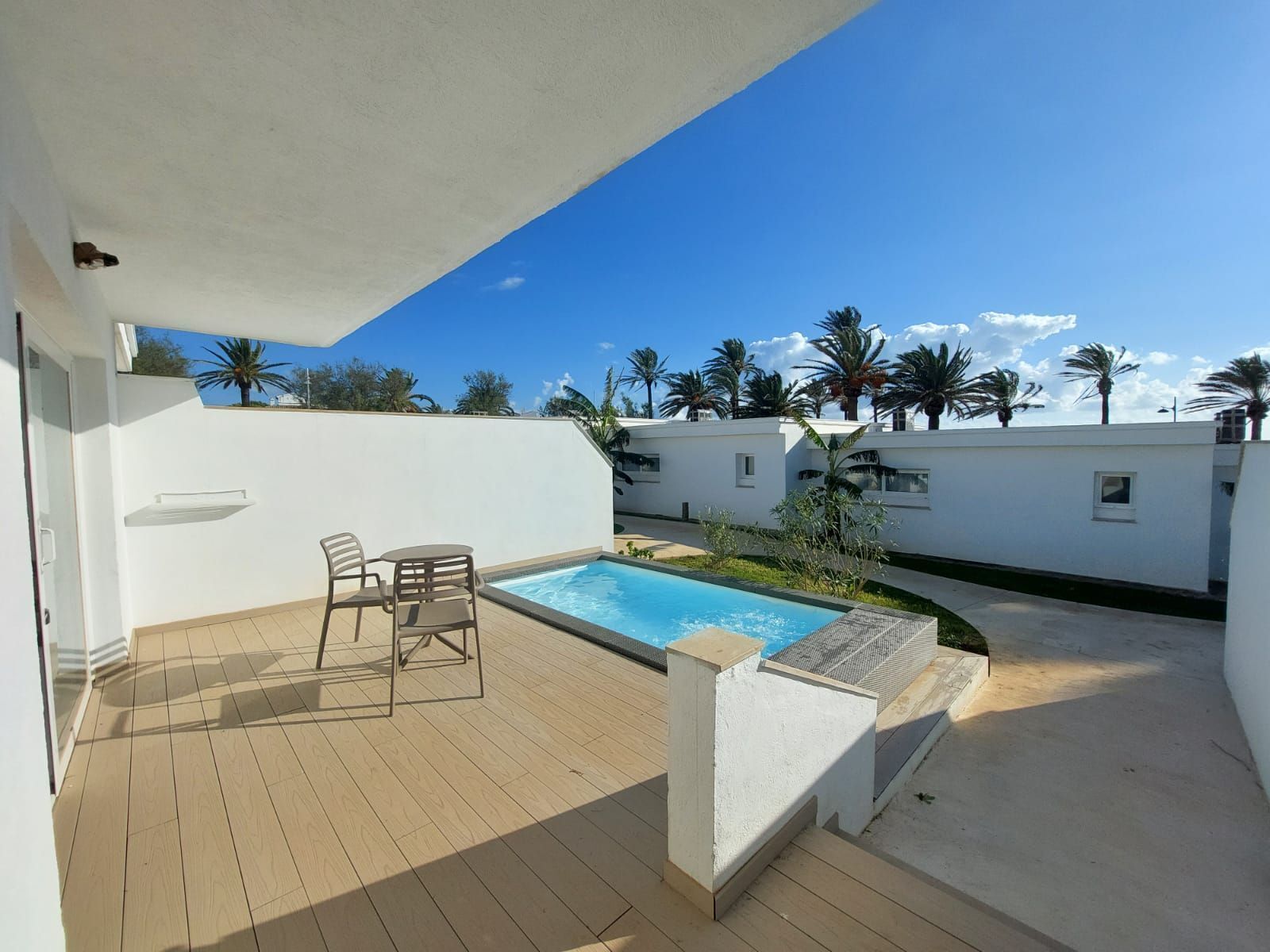 A rooftop pool area with a wooden deck, a small table and chairs, and a view of palm trees and a clear sky.