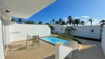 A rooftop pool area with a wooden deck, a small table and chairs, and a view of palm trees and a clear sky.