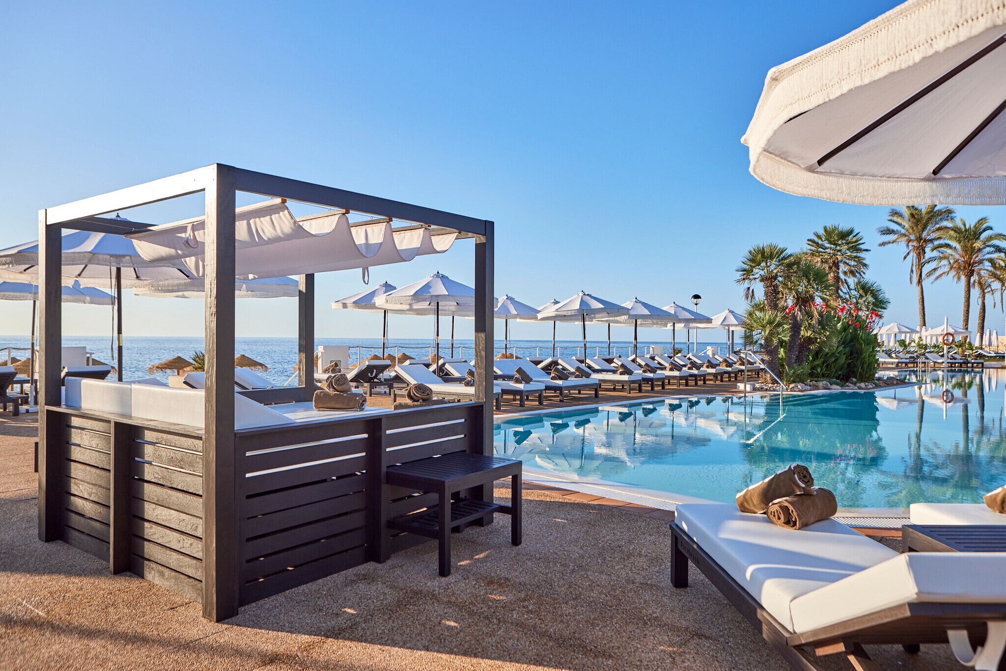 A poolside area with lounge chairs and umbrellas, a wooden structure with a bench, and a view of the sea.