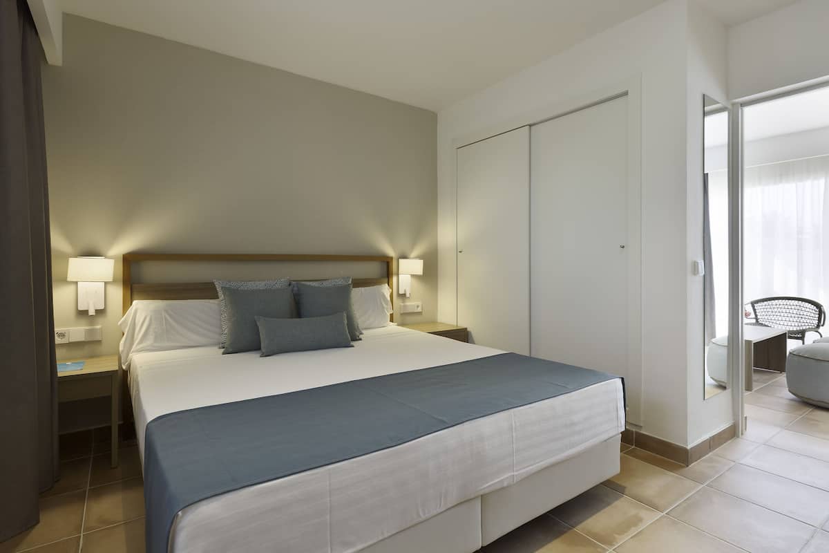 Une chambre d’hôtel avec un grand lit, des tables de chevet avec des lampes, une armoire et une chaise près d’une fenêtre.