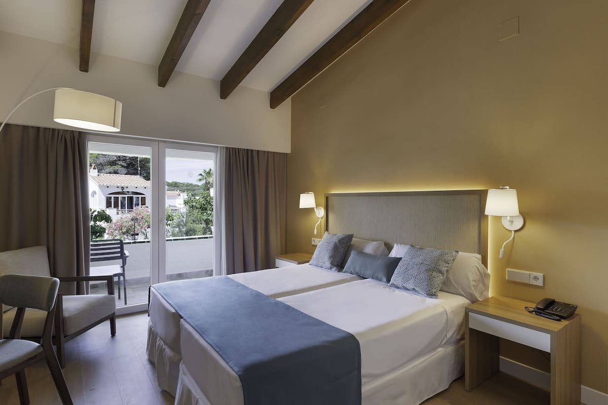 Une chambre d’hôtel avec deux lits, un bureau, une chaise et un balcon offrant une vue sur les bâtiments et la verdure.
