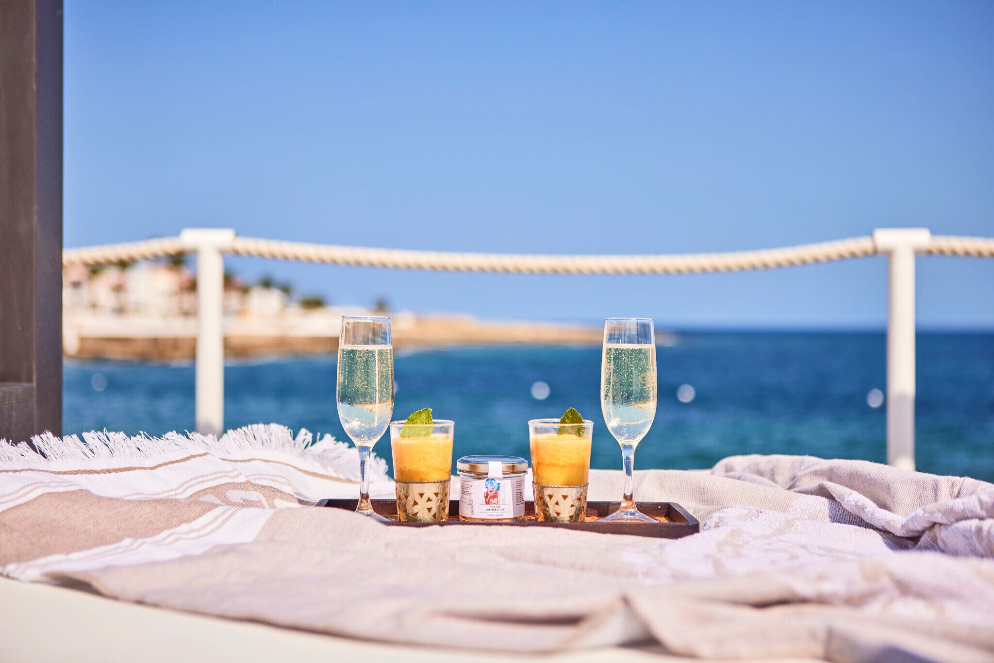 A beachside view with a table set with two glasses of a yellow drink, a small jar, and a bottle, overlooking the sea.