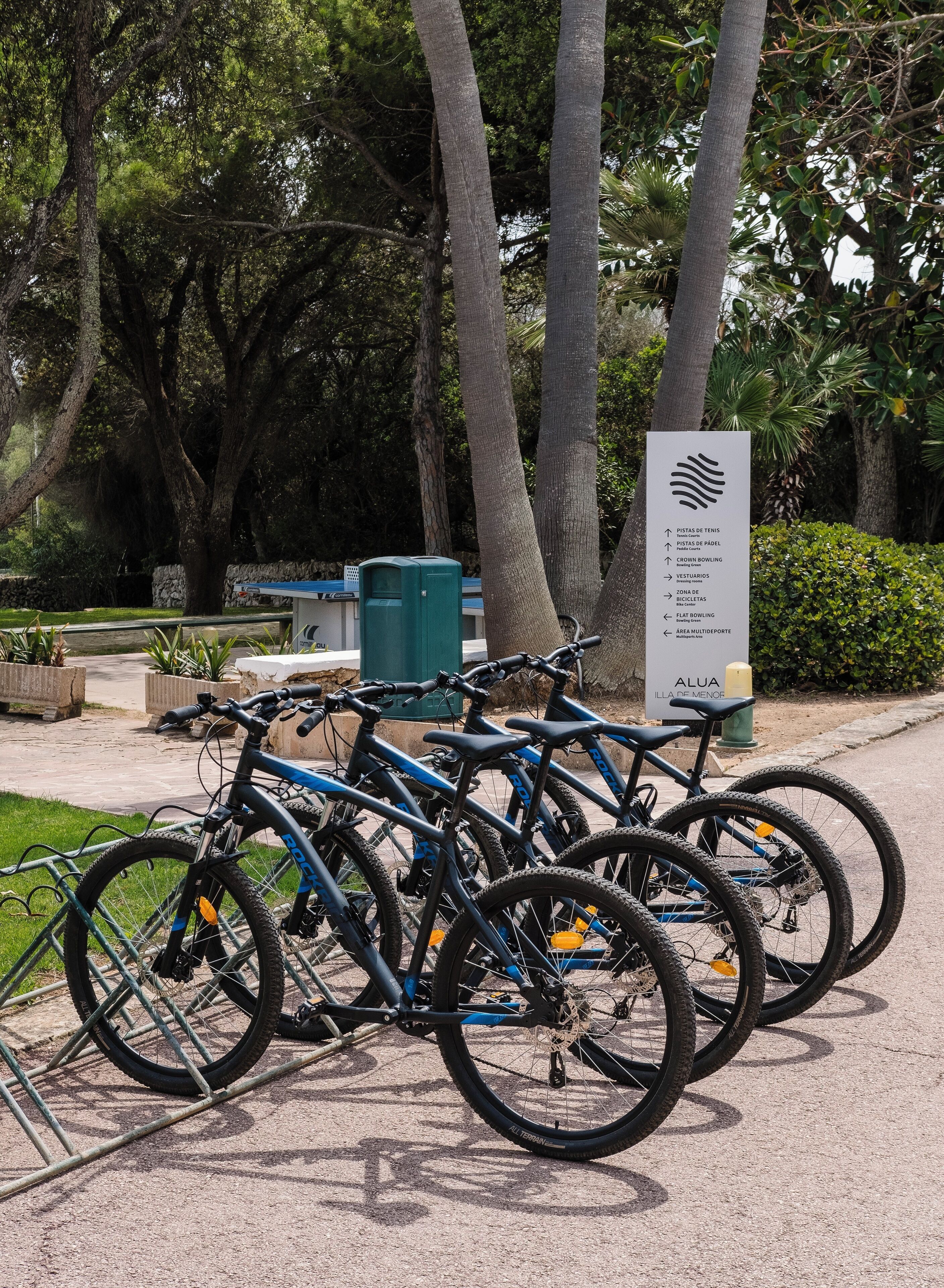 Una fila de bicicletas estacionadas en un área designada, con un cartel que indica servicios como canchas de tenis, un área de picnic y una zona para bicicletas.
