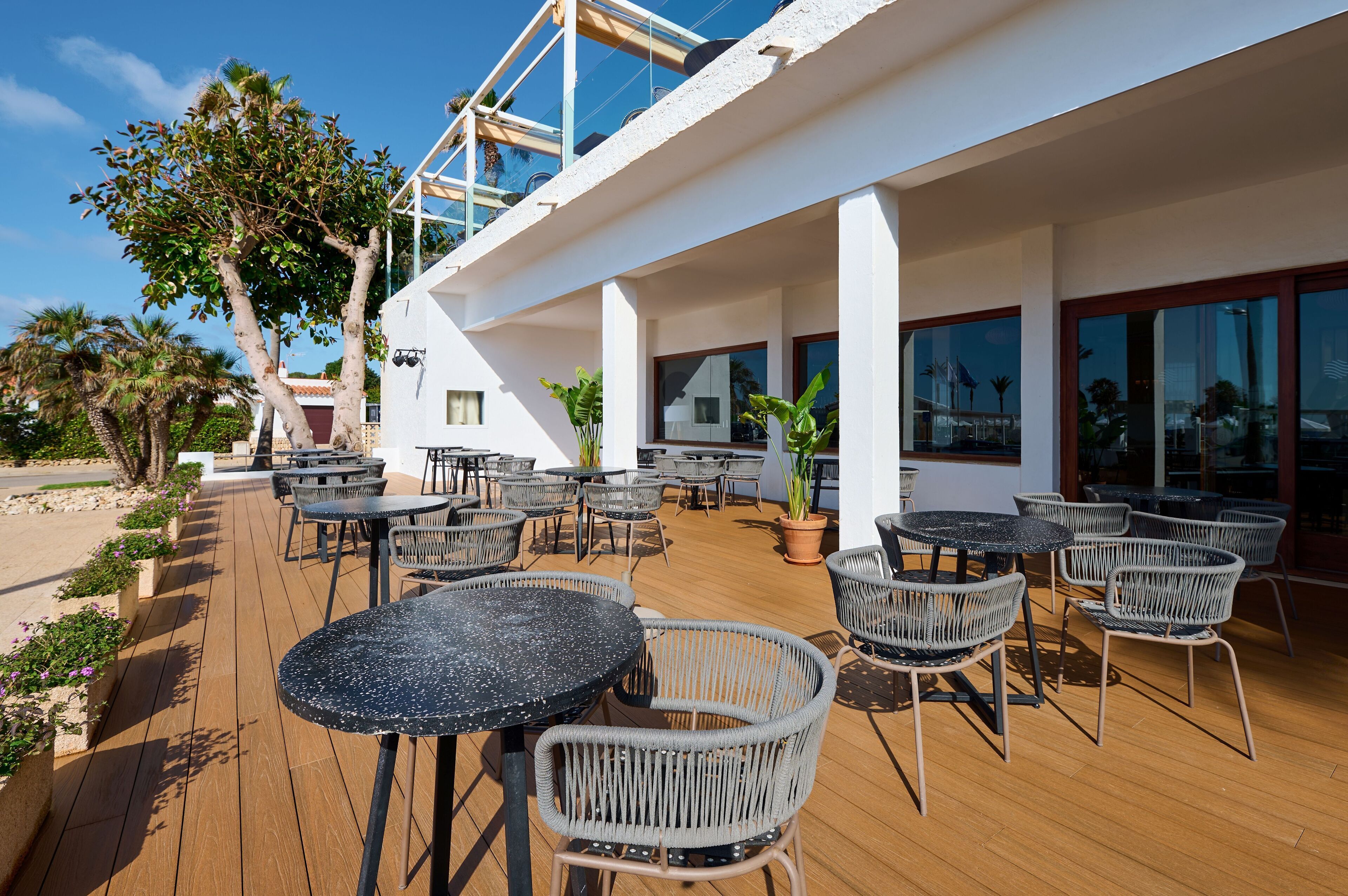 An outdoor patio with round tables and wicker chairs, a wooden deck, and a white building with large windows.