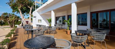 Un patio al aire libre con mesas redondas y sillas de mimbre, una terraza de madera y un edificio blanco con ventanales grandes.