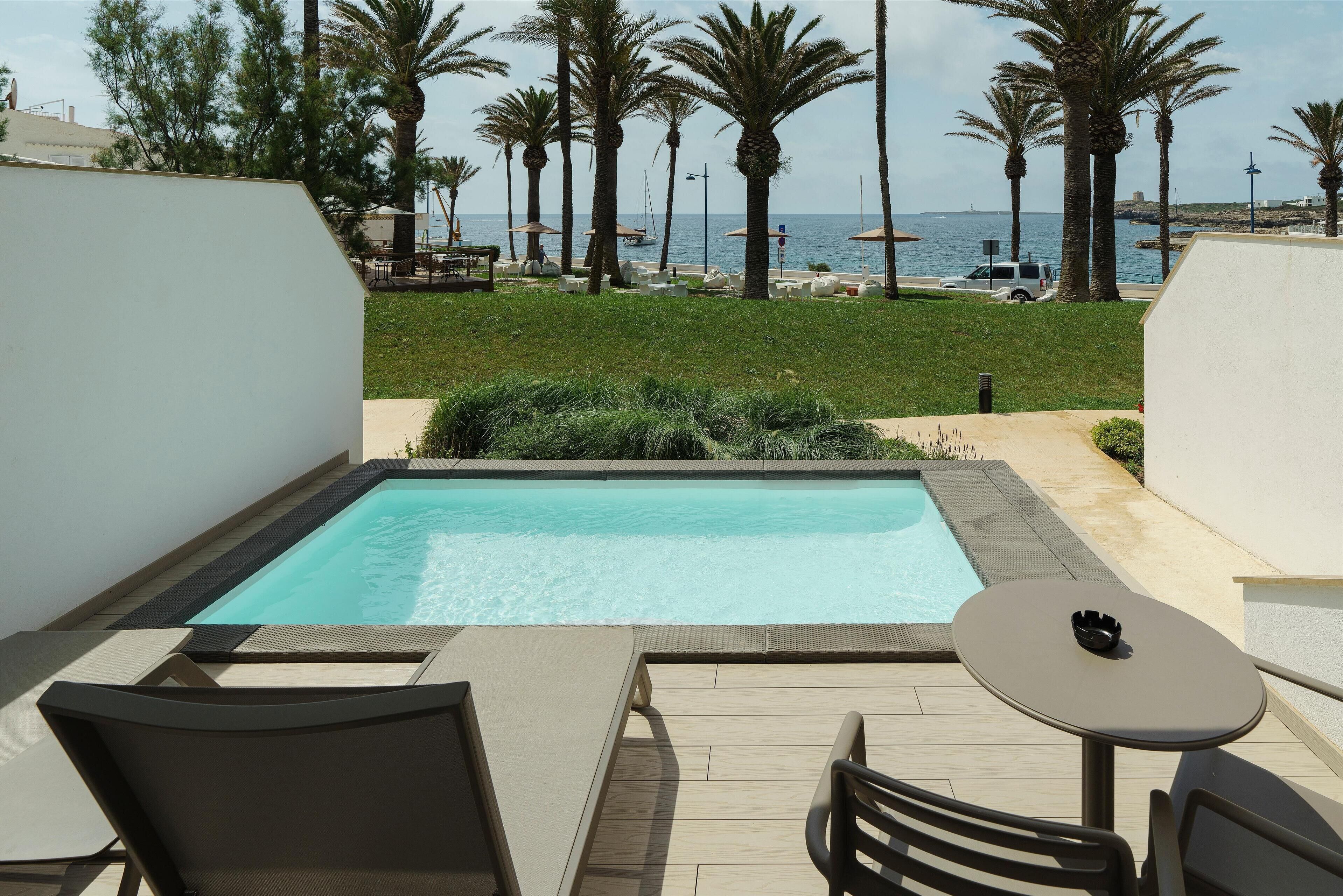 Zona de piscina con vista al mar, rodeada de palmeras y un edificio blanco.