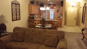 A living room with a brown patterned sofa, a dining table set for four, wooden cabinets, and a kitchen area with a window and a door. - Royal Links (Branson)