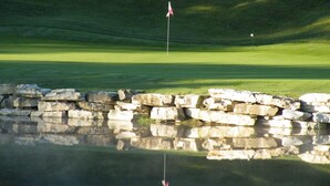 A golf course with a flagstick in the center, a pond reflecting the flagstick, and a stone wall in the foreground. - Royal Links (Branson)
