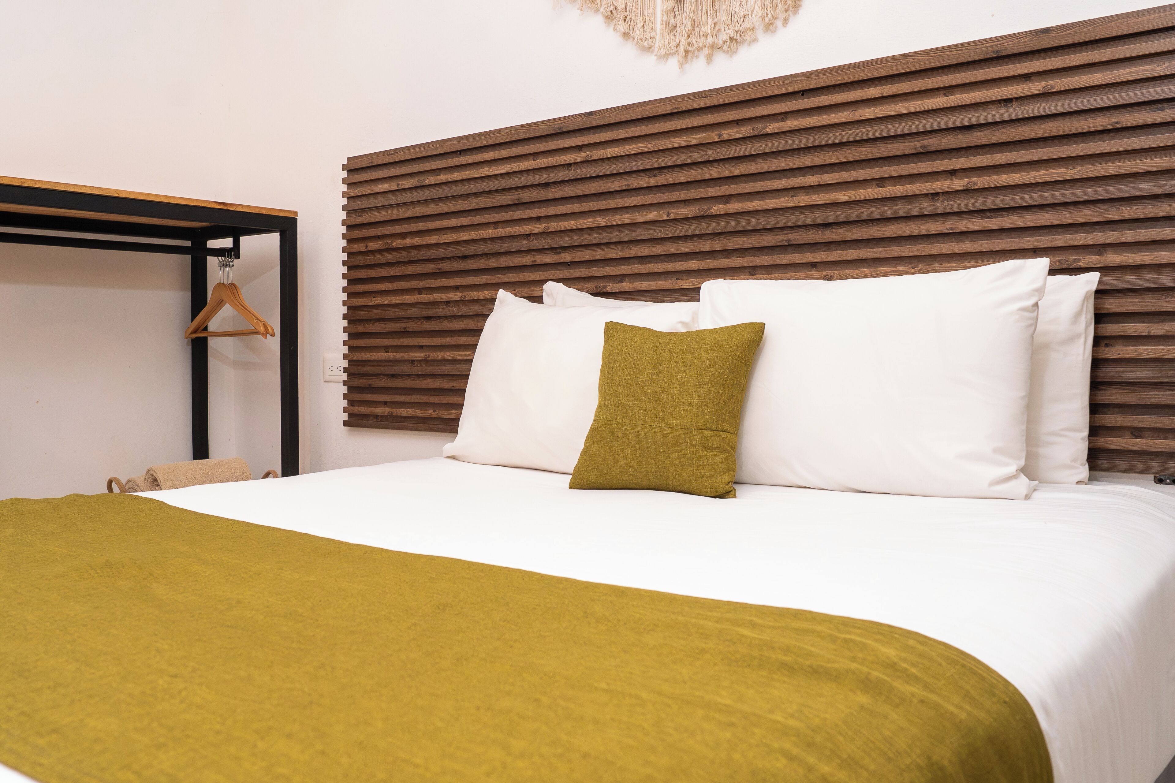 A neatly made bed with white and olive green bedding, a wooden headboard, and a simple wooden hanger on the left.