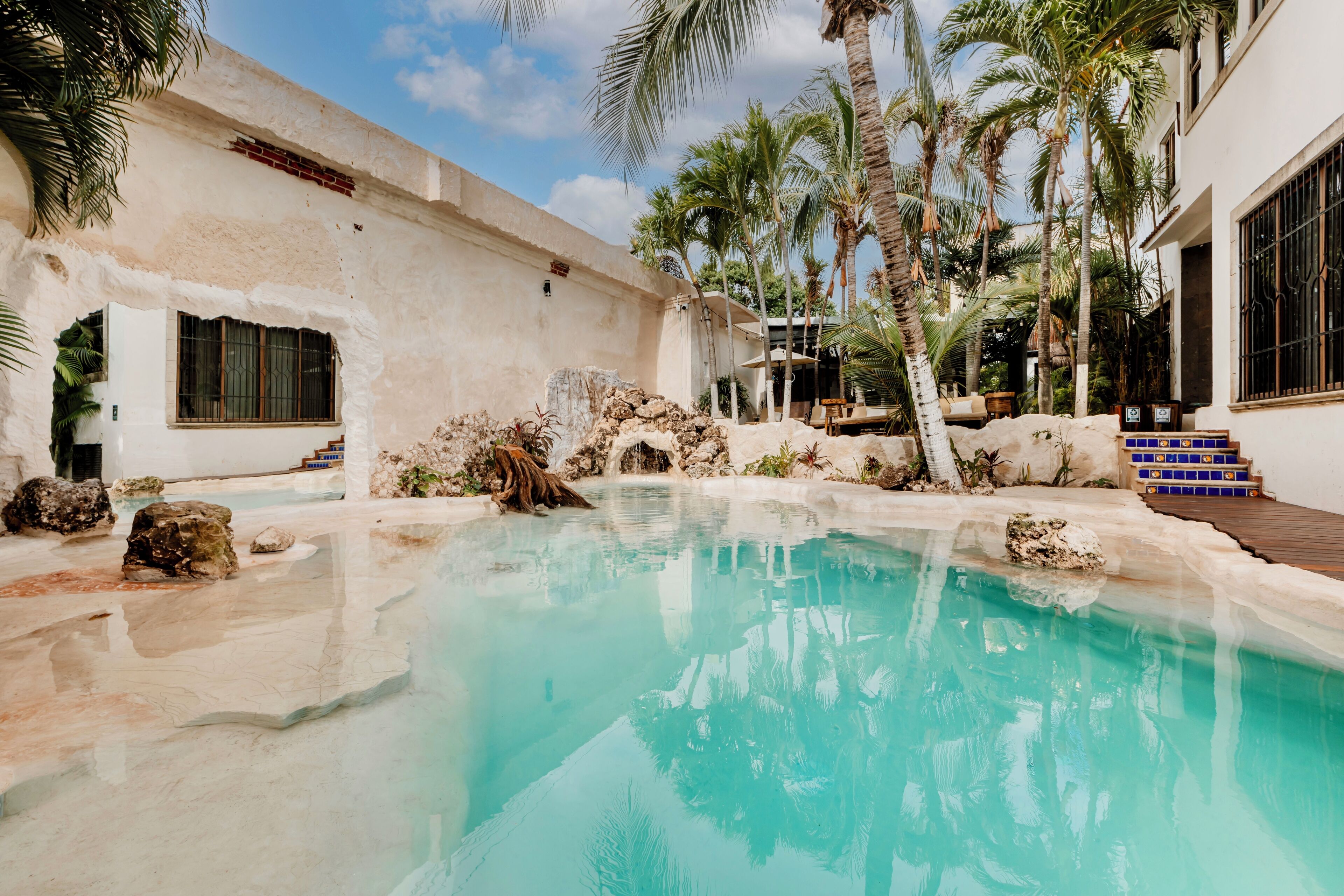 A swimming pool with clear water, surrounded by rocks and palm trees, and a building with large windows and a white exterior.