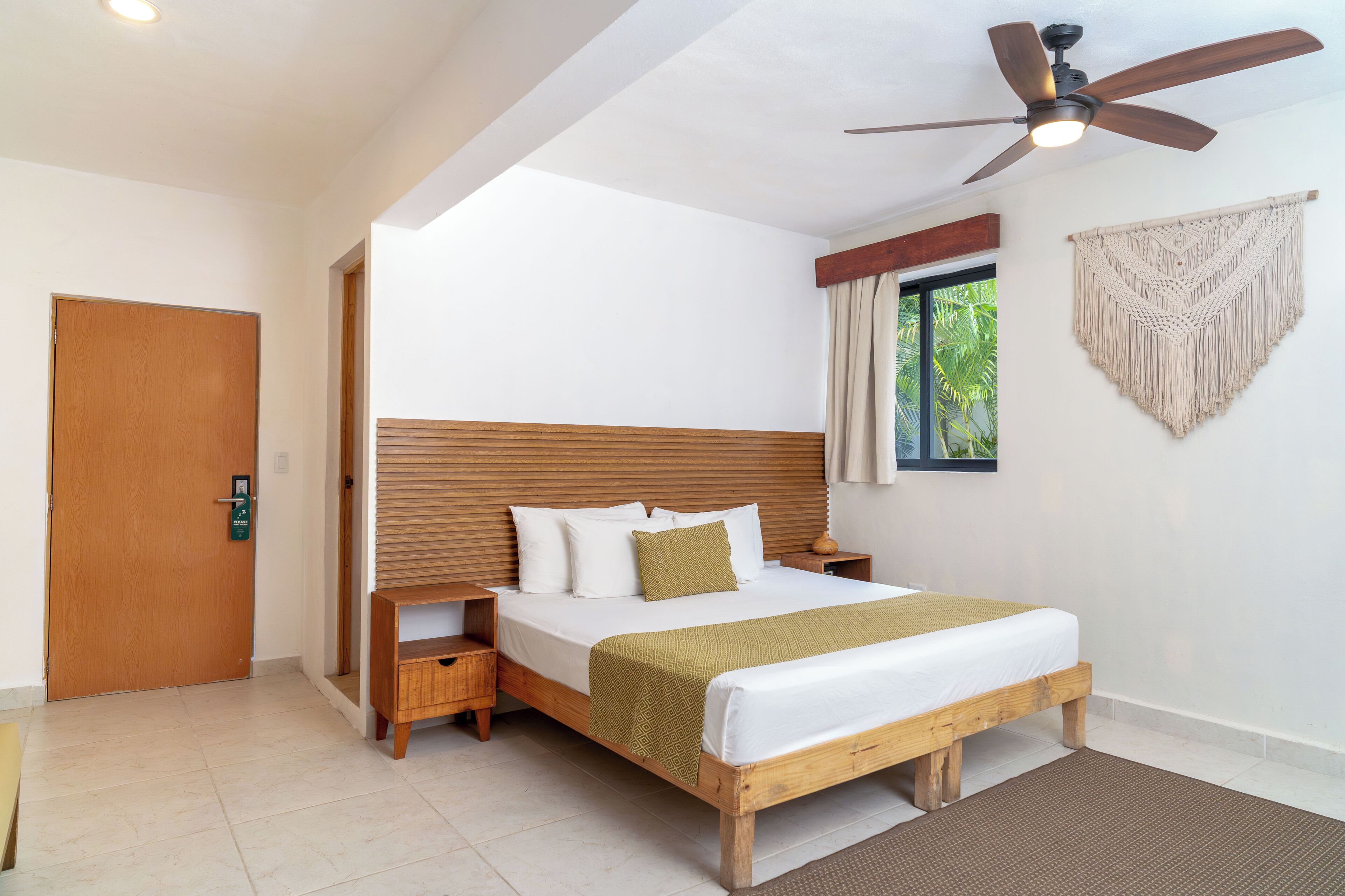Een hotelkamer met een houten bed, een nachtkastje, een plafondventilator, een raam met gordijnen en een wanddecoratie van macramé.