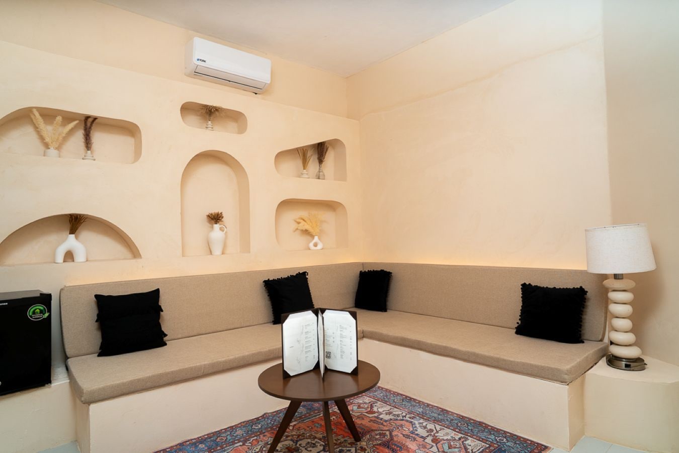 Une pièce avec un canapé beige, des coussins noirs, une petite table sur laquelle se trouve un livre, une lampe et des vases décoratifs accrochés au mur.