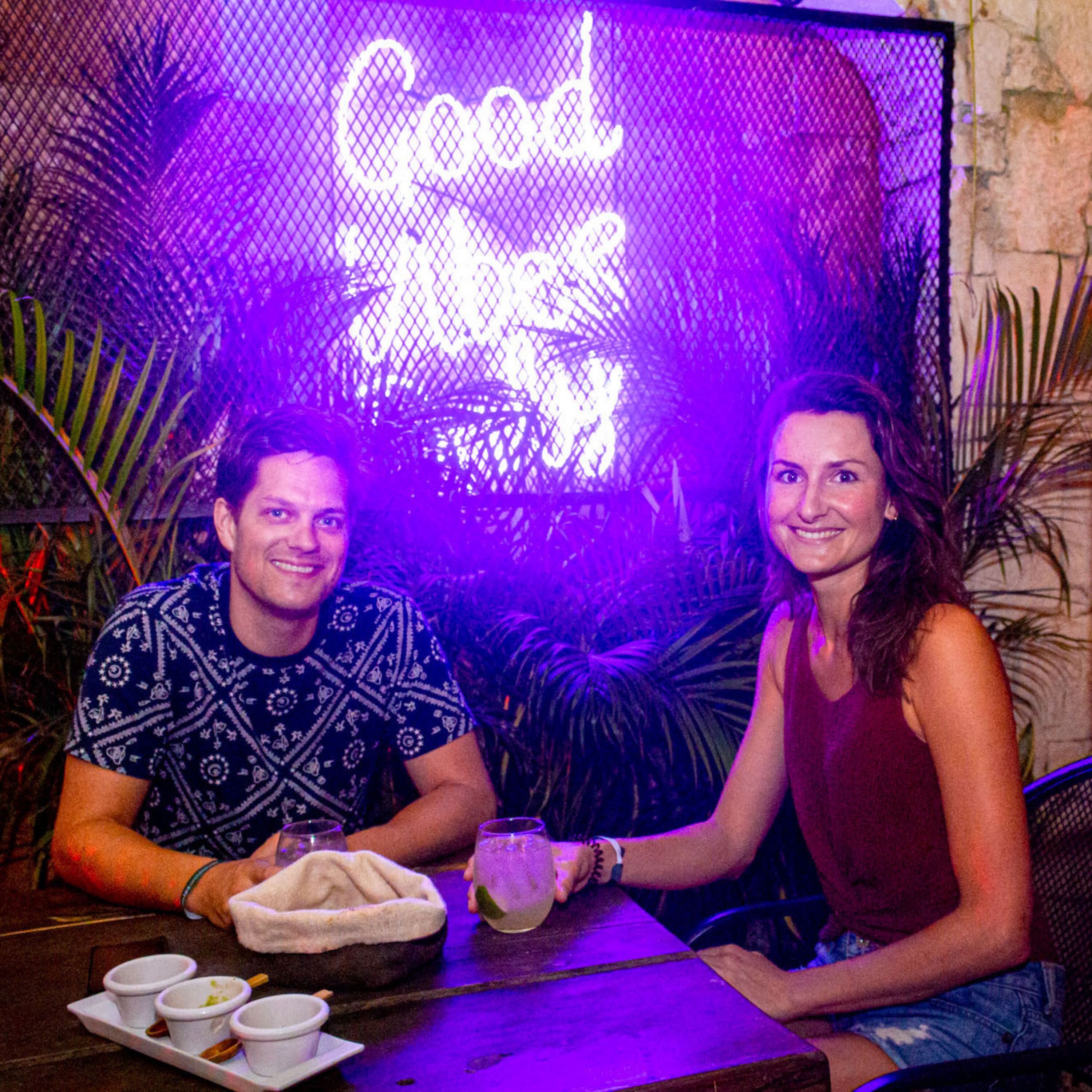 Two people seated at a table with a neon sign in the background.