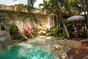 Una piscina con cascada, rodeada de plantas tropicales y una terraza de madera.