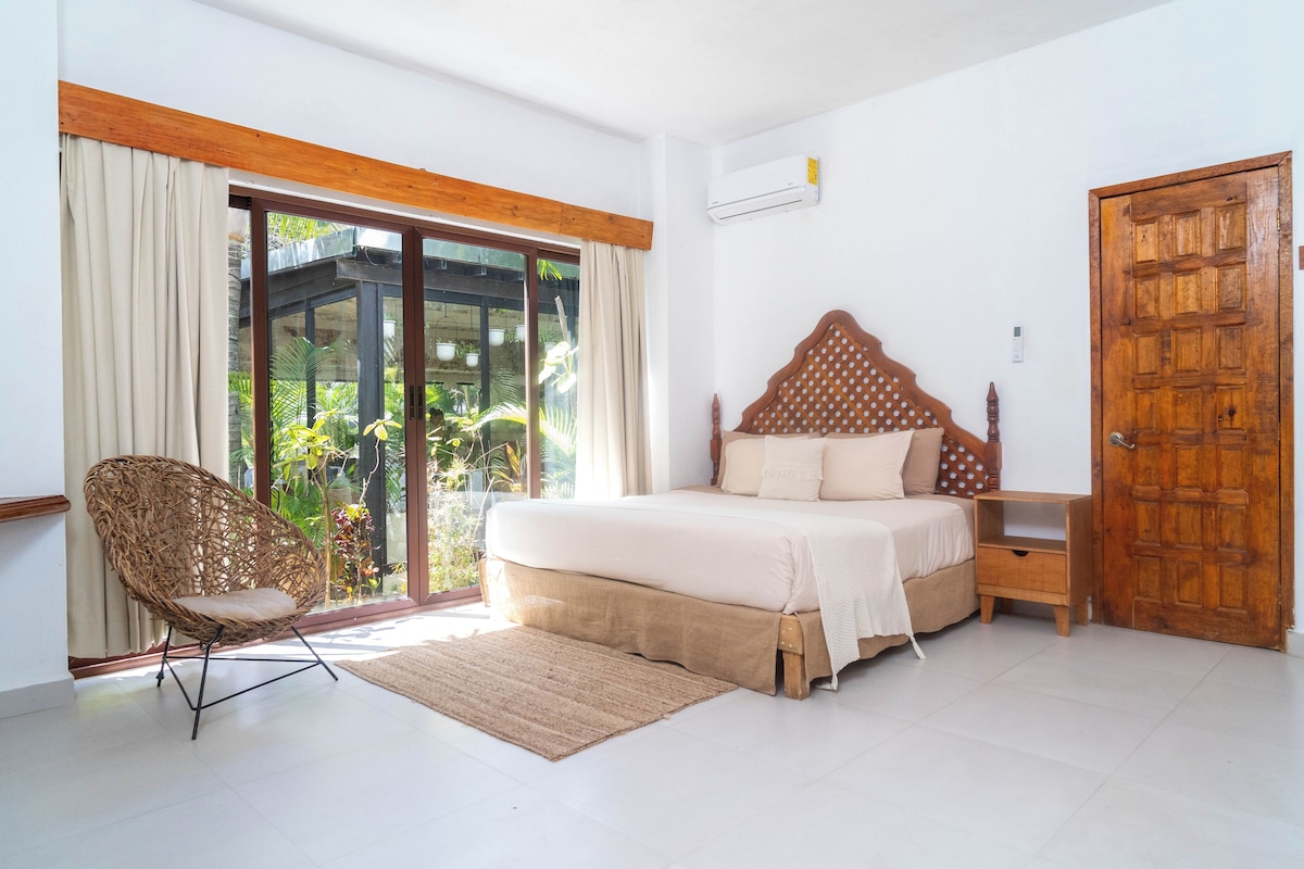 A bedroom with a bed, a wooden door, a window with curtains, and a wicker chair.