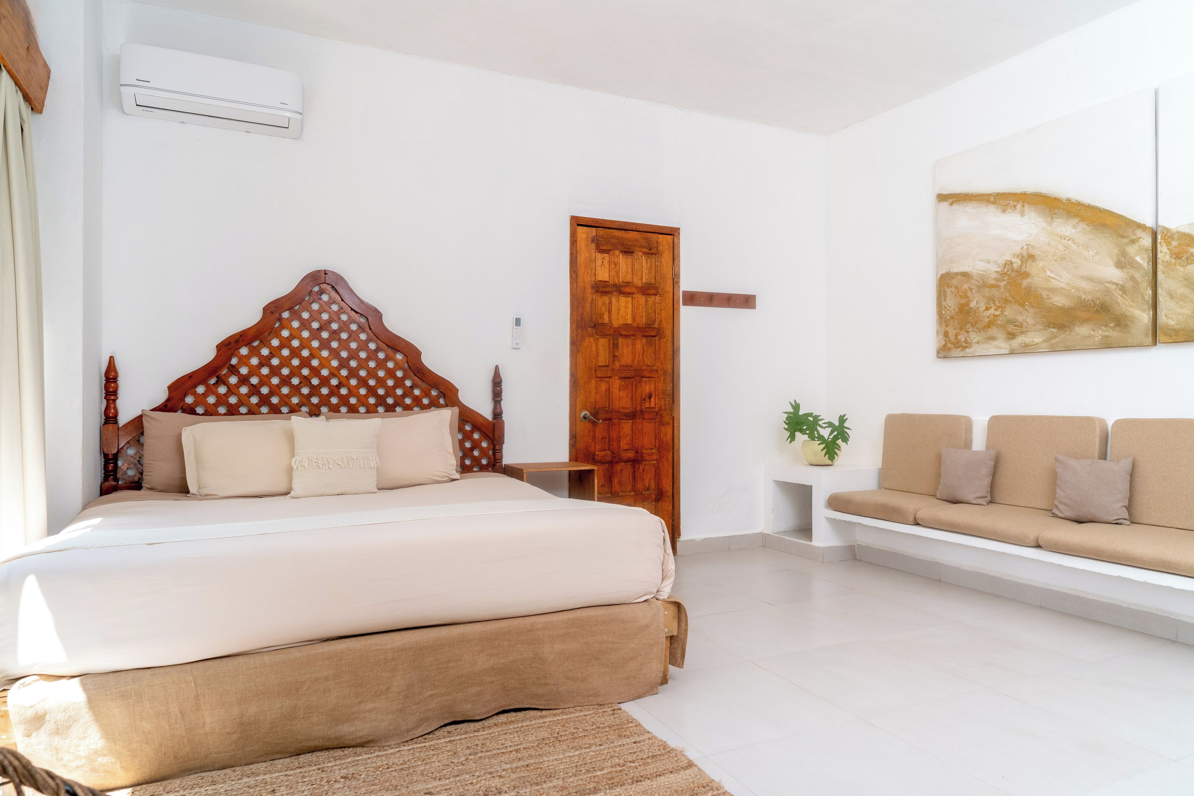 A bedroom with a bed, a wooden headboard, a wooden door, a white sofa, and a wall-mounted air conditioner.