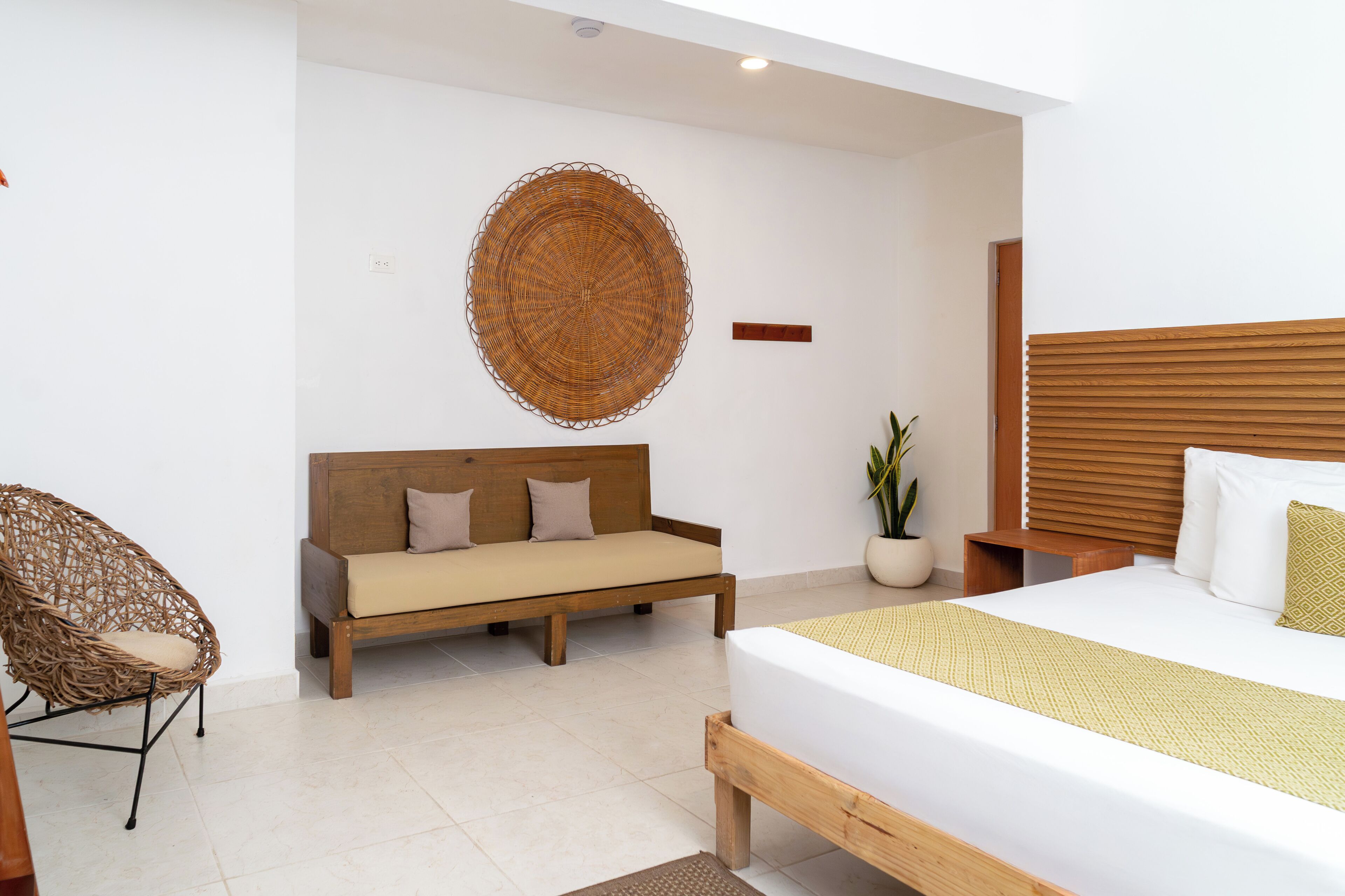 Quarto moderno com cama de madeira, sofá, poltrona de vime e planta em vaso.