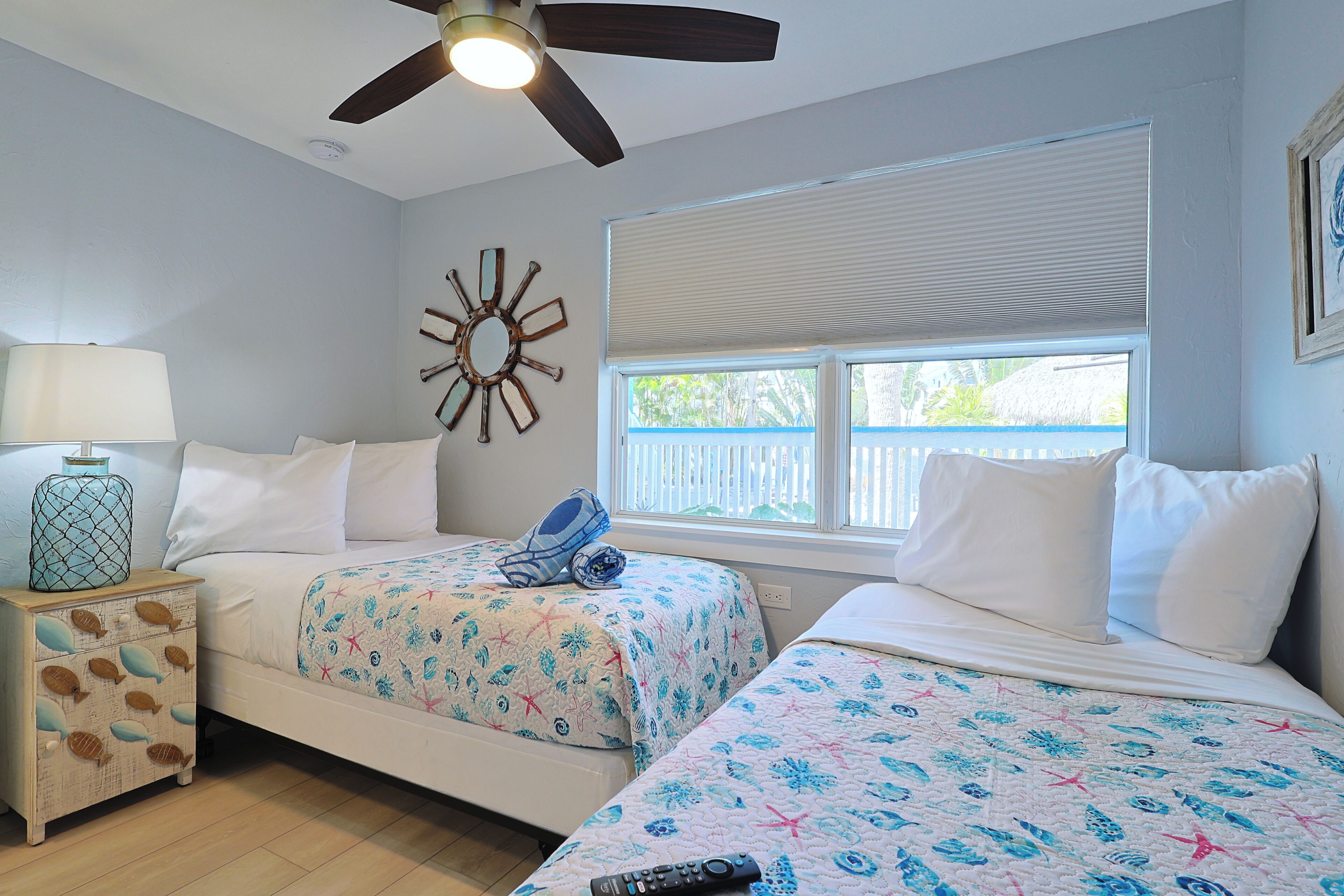 A bedroom with two beds, a ceiling fan, a window with blinds, a nightstand with a lamp, and a decorative wall piece.