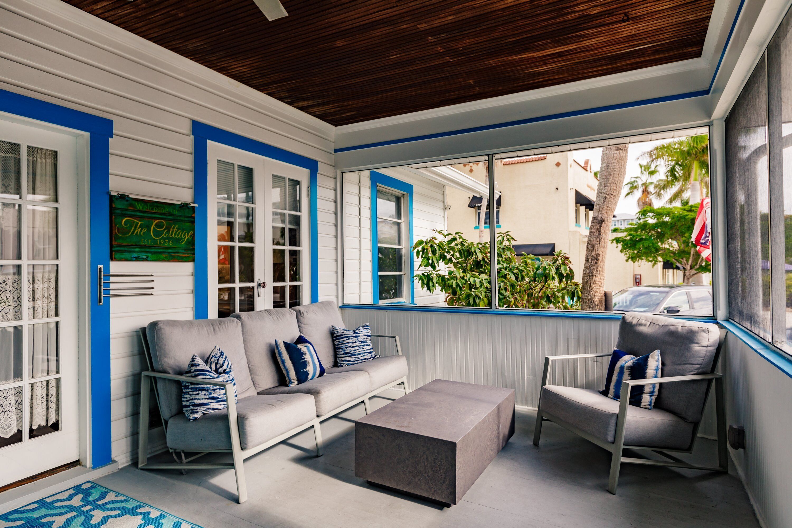 A covered porch with a gray sofa, a gray armchair, and a small table. There is a sign that reads "The Cottage" and a window with blue trim.