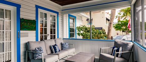 A covered porch with a gray sofa, a gray armchair, and a small table. There is a sign that reads "The Cottage" and a window with blue trim.
