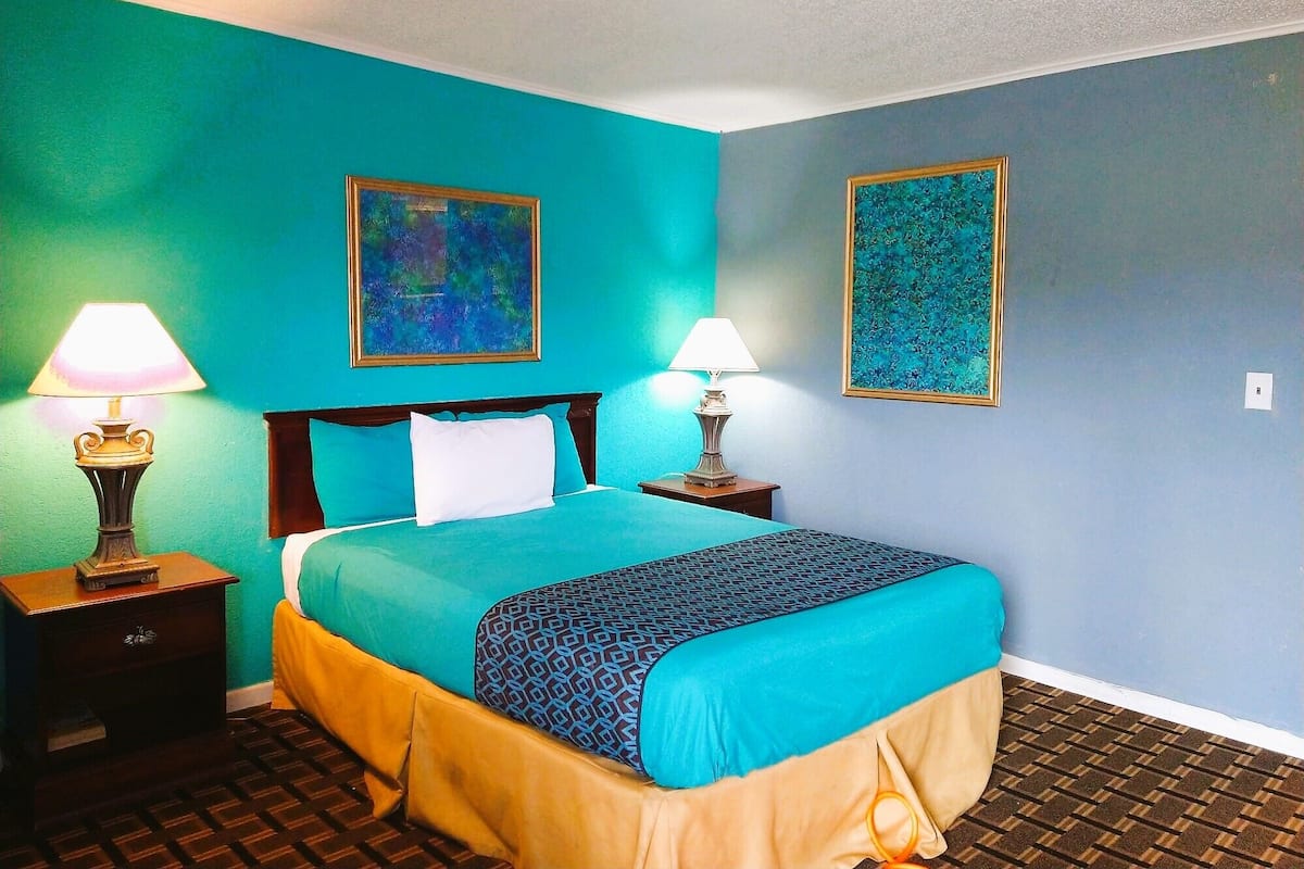 Une pièce aux murs de couleur turquoise, un lit recouvert d’une couvre-lit à motifs, deux lampes de chevet et deux tableaux abstraits encadrés.