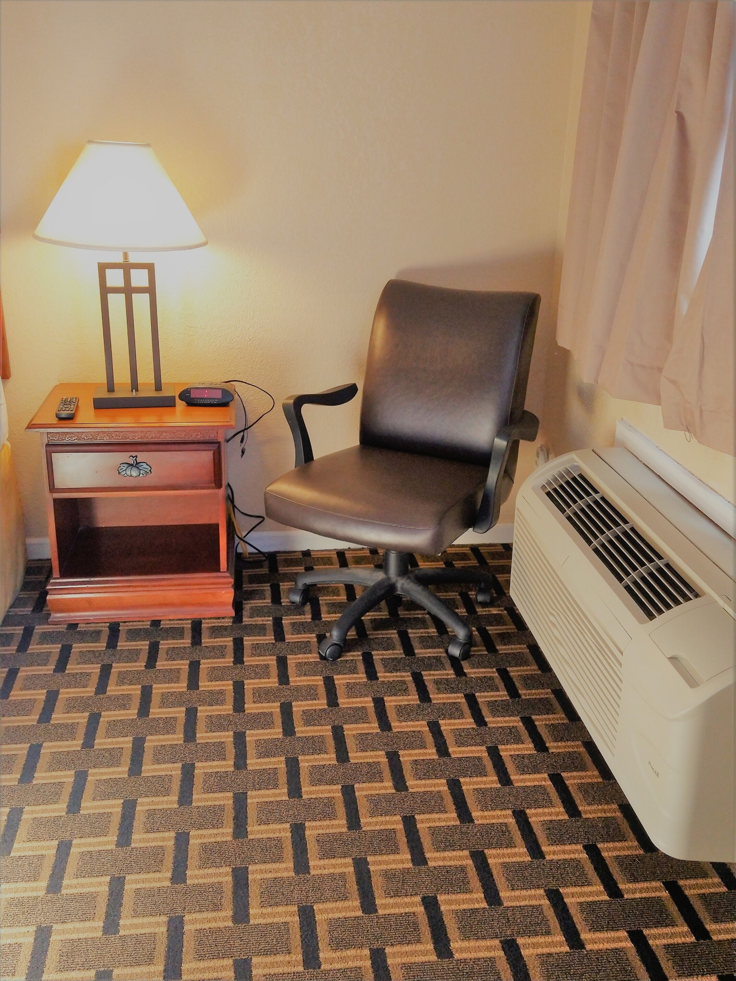 A hotel room with a single office chair, a desk with a lamp, a small drawer unit, and a portable heater.
