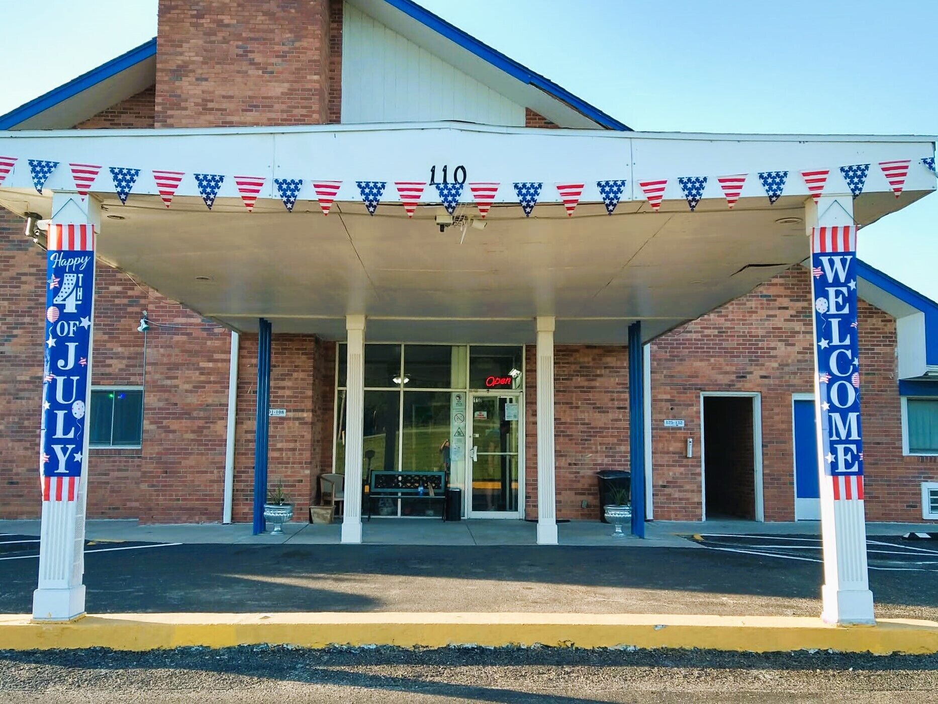 A building entrance with a covered area, a 'Welcome' sign, and a 'Happy 4th of July' banner.