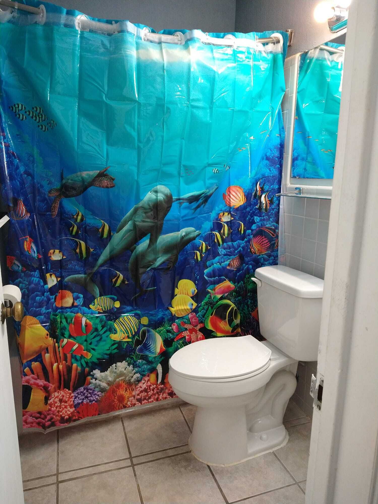 A bathroom with a blue aquarium-themed shower curtain, a white toilet, and a small window above the toilet.